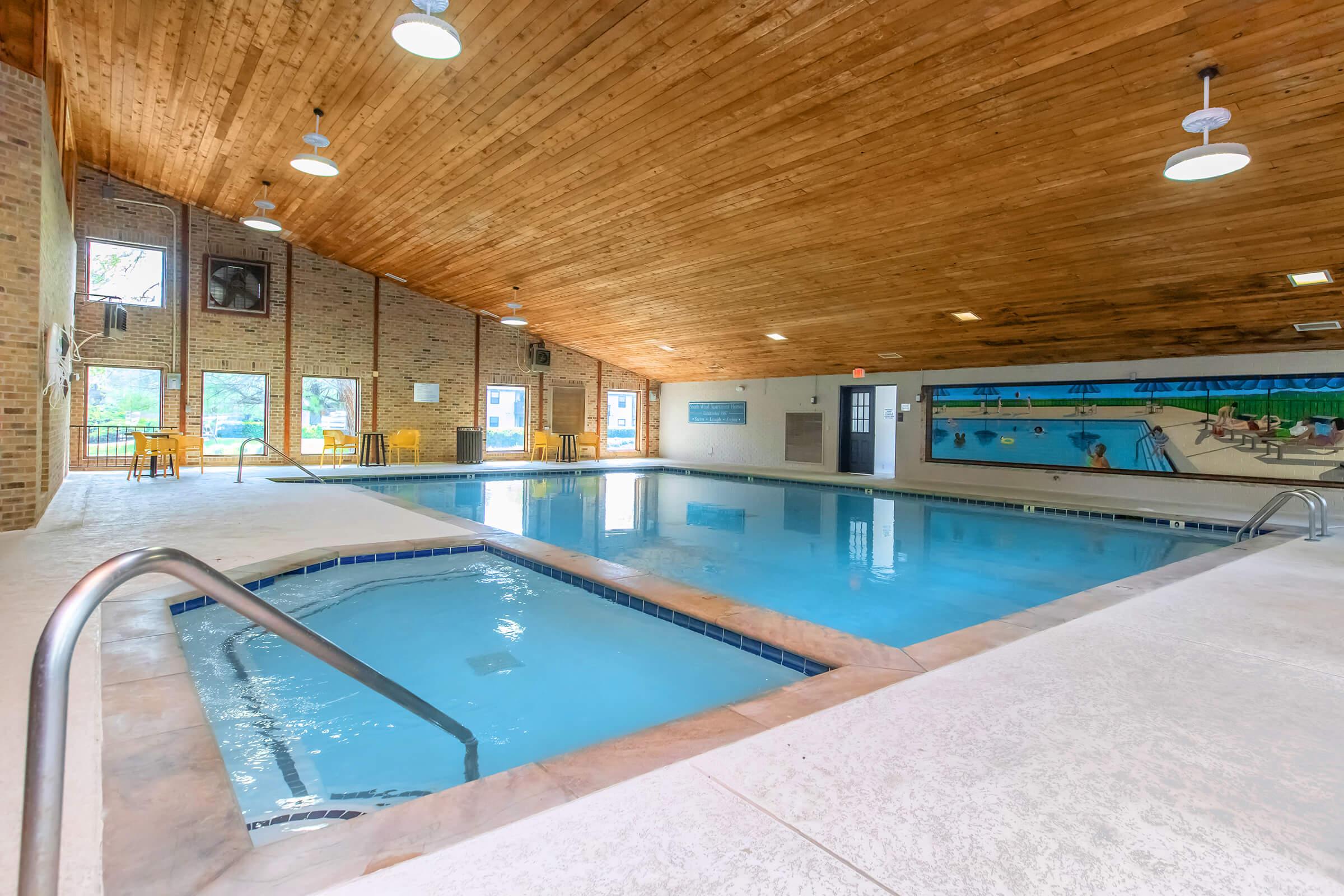 Indoor swimming pool area featuring a spacious layout with a wooden ceiling and large windows allowing natural light. One side has a swimming pool with clean water, while the other displays a mural of swimmers. There are chairs and tables along the poolside, creating a relaxing atmosphere.