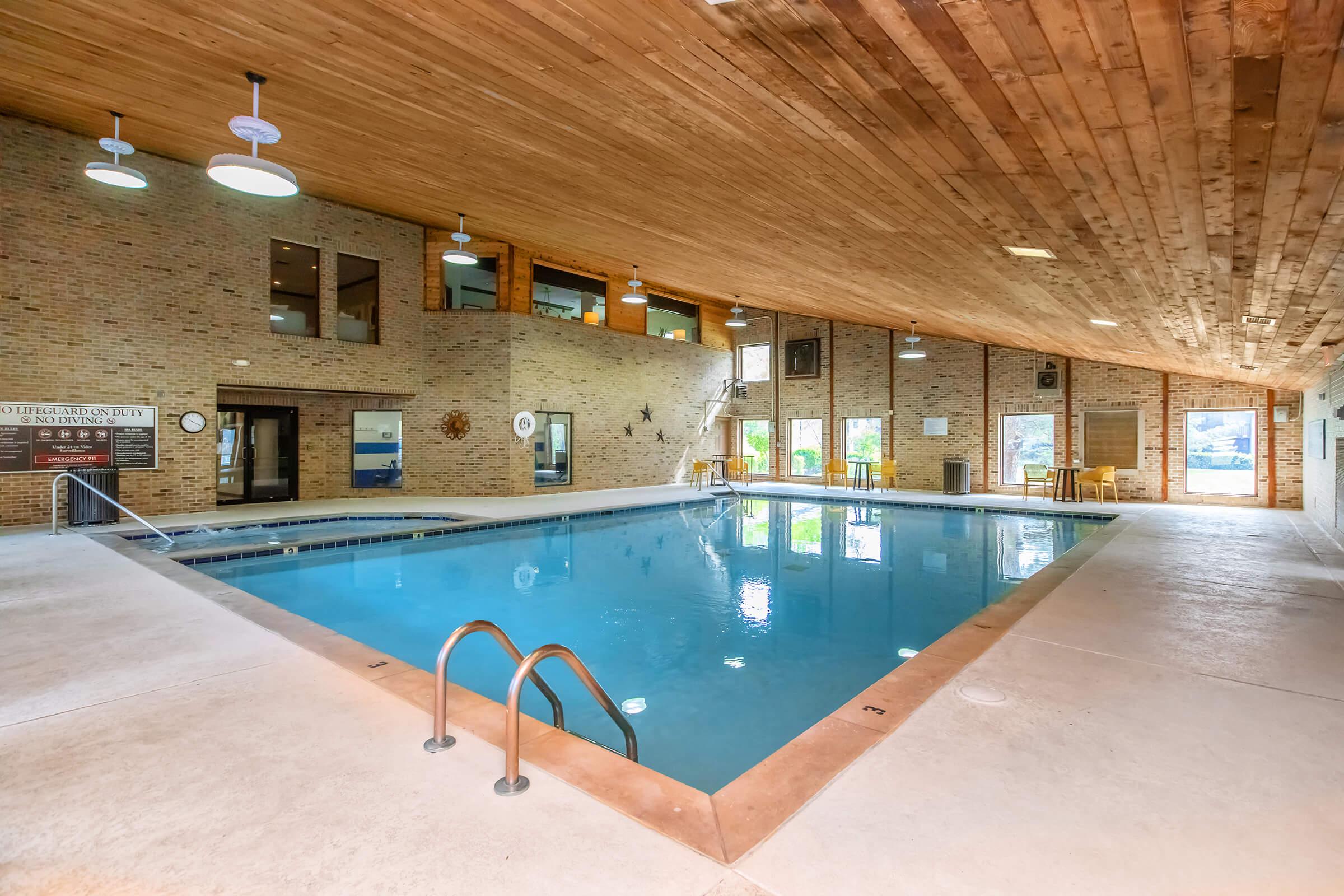 Indoor swimming pool with clear blue water, surrounded by a light-colored stone deck. The ceiling features wooden beams, and large windows allow natural light to fill the space. There are chairs and tables near the pool area, creating a relaxing atmosphere.