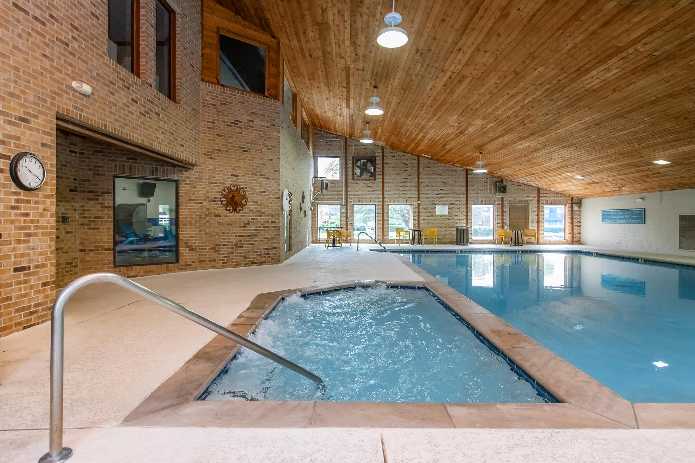 Indoor swimming pool area featuring a hot tub, wood-paneled ceiling, and brick walls. The pool is unoccupied and surrounded by large windows that allow natural light to enter. There are lounge chairs placed around the pool, and a clock is visible on the wall.