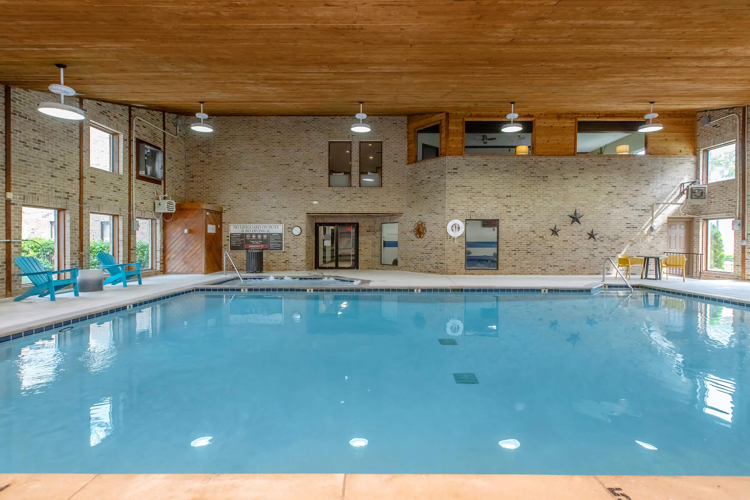 Indoor swimming pool area with clear blue water, surrounded by brick walls and wooden ceiling. Lounge chairs are arranged nearby, with large windows allowing natural light to enter. A diving board and a pool ladder are visible, along with decorative elements like stars on the walls.
