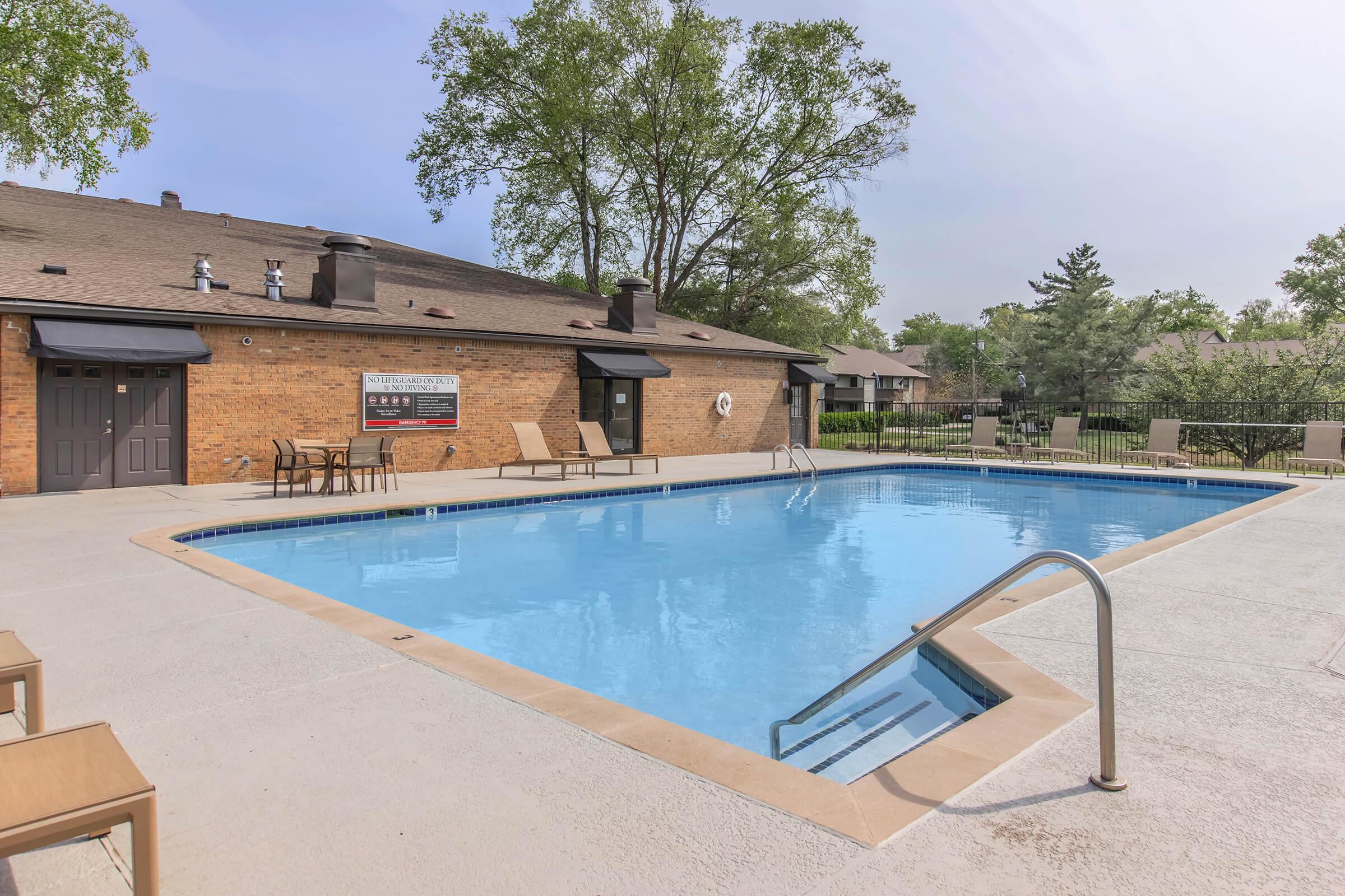 A clear blue swimming pool with a shallow entry, surrounded by a concrete deck. Several lounge chairs are positioned nearby, and a brick building with dark windows is seen in the background. Lush green trees and a well-maintained landscape create a serene atmosphere.
