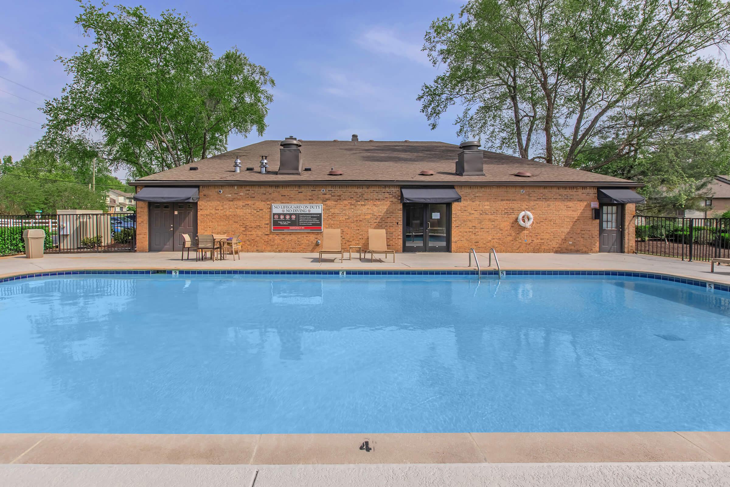 A clear blue swimming pool surrounded by a concrete deck, with several lounge chairs positioned nearby. In the background, there is a brick building with multiple windows and a sign. Lush green trees provide shade around the area, creating a relaxing atmosphere.