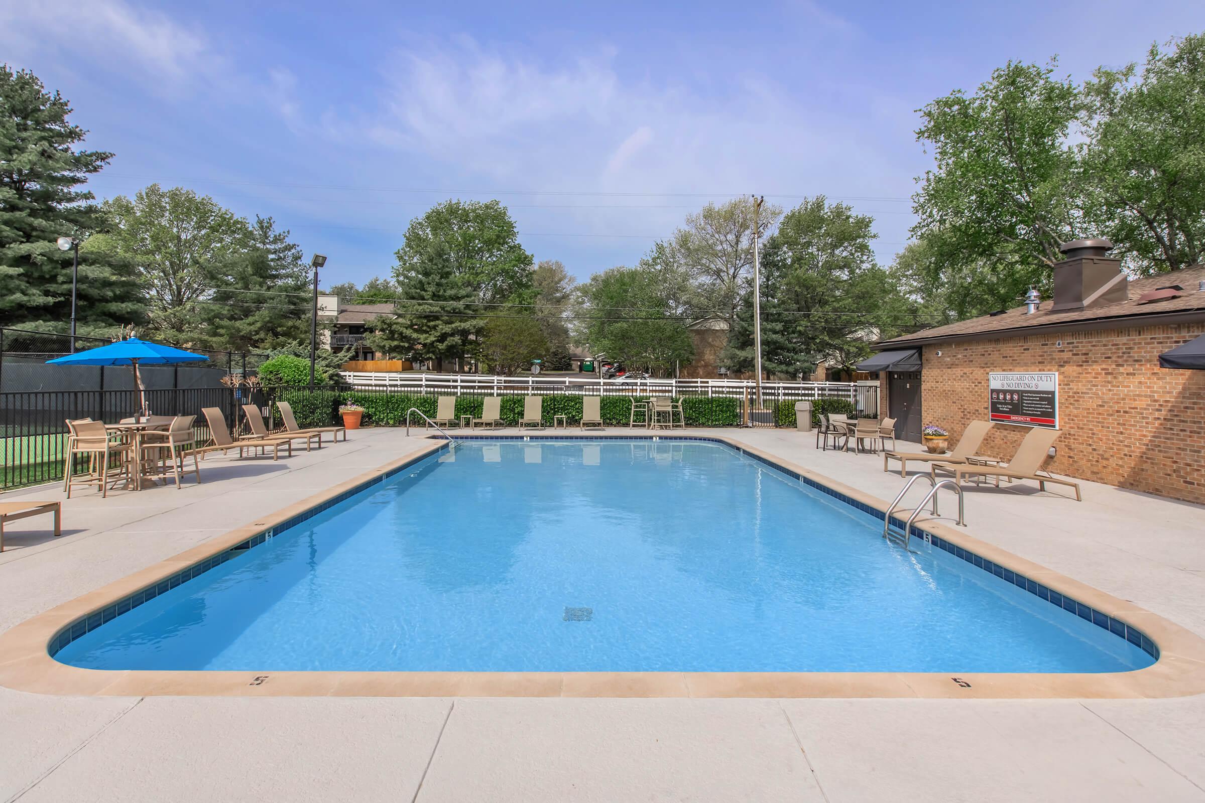 A serene outdoor pool area featuring a clear blue pool surrounded by lounge chairs and umbrellas. Lush greenery and trees are visible in the background, along with a brick building that adds warmth to the setting. The area is inviting, perfect for relaxation or leisure activities.