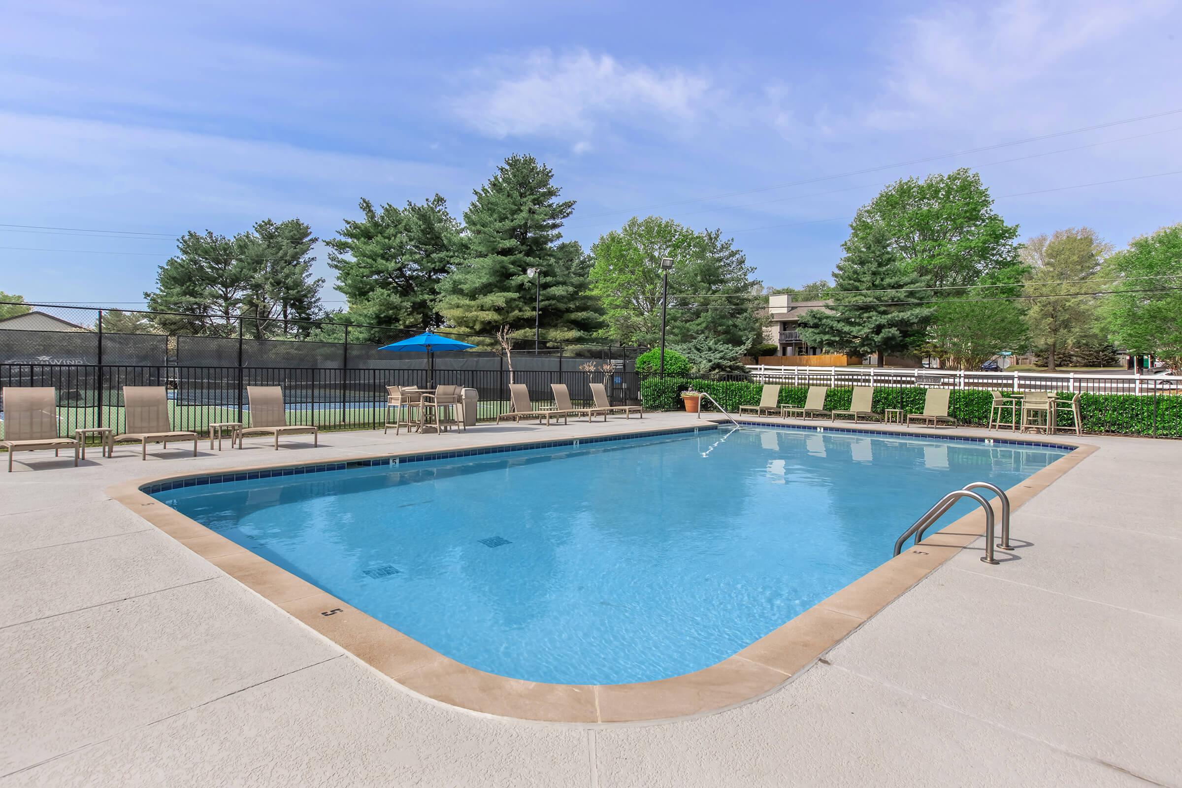 A clear blue swimming pool surrounded by lounge chairs and green trees. A shaded area with a blue umbrella can be seen in the background, along with a well-maintained lawn area. The sky is partly cloudy, creating a bright and inviting atmosphere.