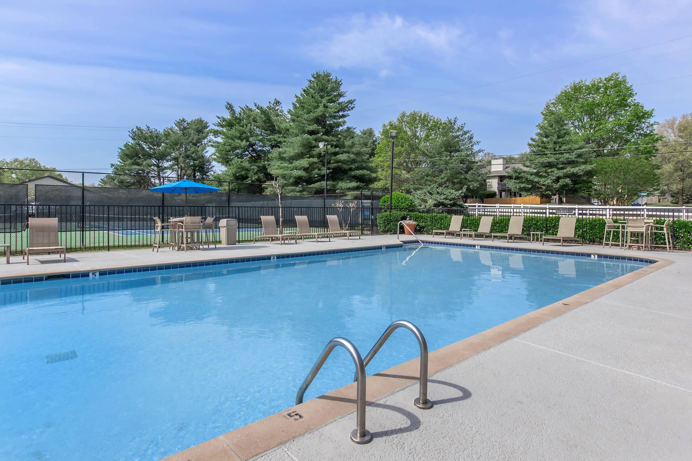 A clear swimming pool with ladders, surrounded by lounge chairs and greenery. In the background, there are trees and a blue umbrella, with a fence enclosing the area under a bright sky.