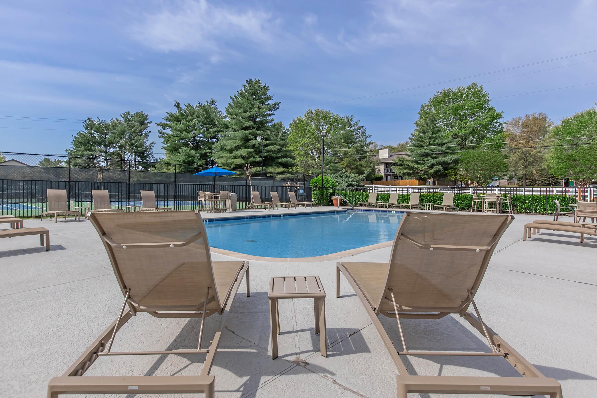 Outdoor pool area featuring two lounge chairs and a small table. Surrounded by greenery and trees, the pool is clear and inviting, with additional seating and umbrellas in the background under a bright blue sky. Perfect for relaxation and leisure.