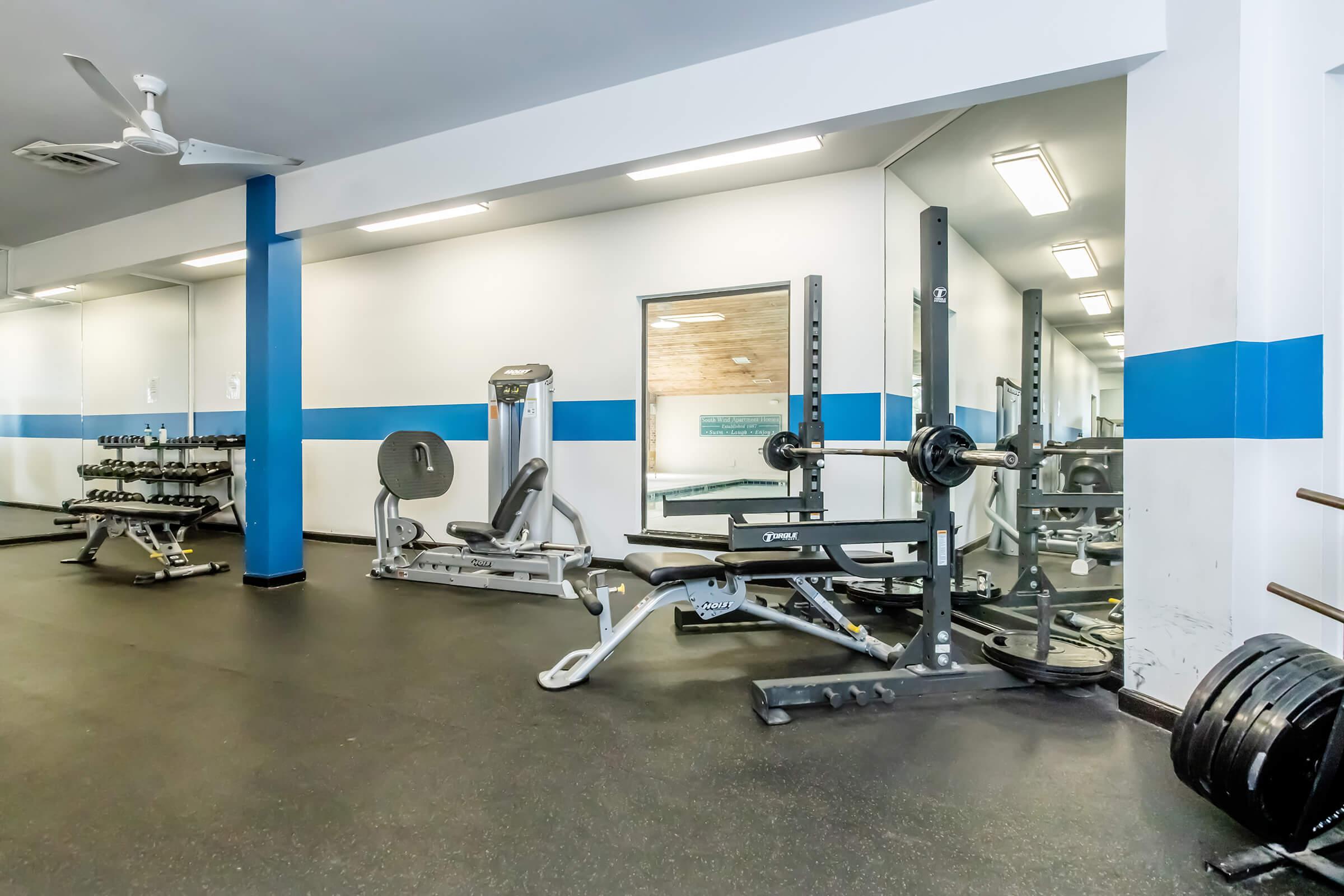 A well-equipped gym featuring various exercise machines, free weights on racks, a barbell with weight plates, and mirrors reflecting the space. The walls are painted with blue accents, and the floor is covered with dark mats, providing a functional workout environment.