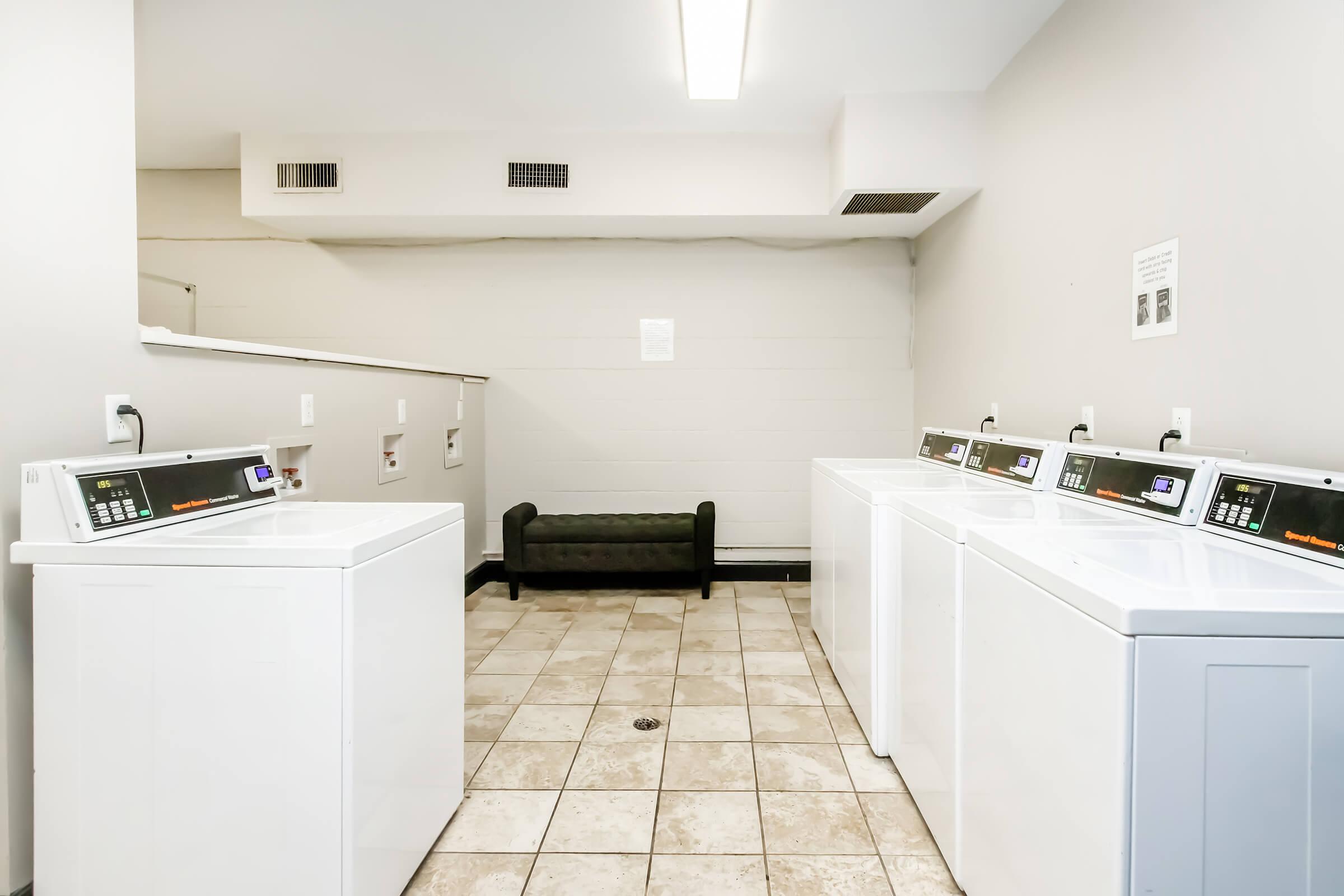 A bright and clean laundry room featuring several white washing machines and dryers lined up against the wall. There is a black bench for seating in the background and tiled flooring, creating a spacious and organized environment. The walls are painted in a light color with minimal decor.