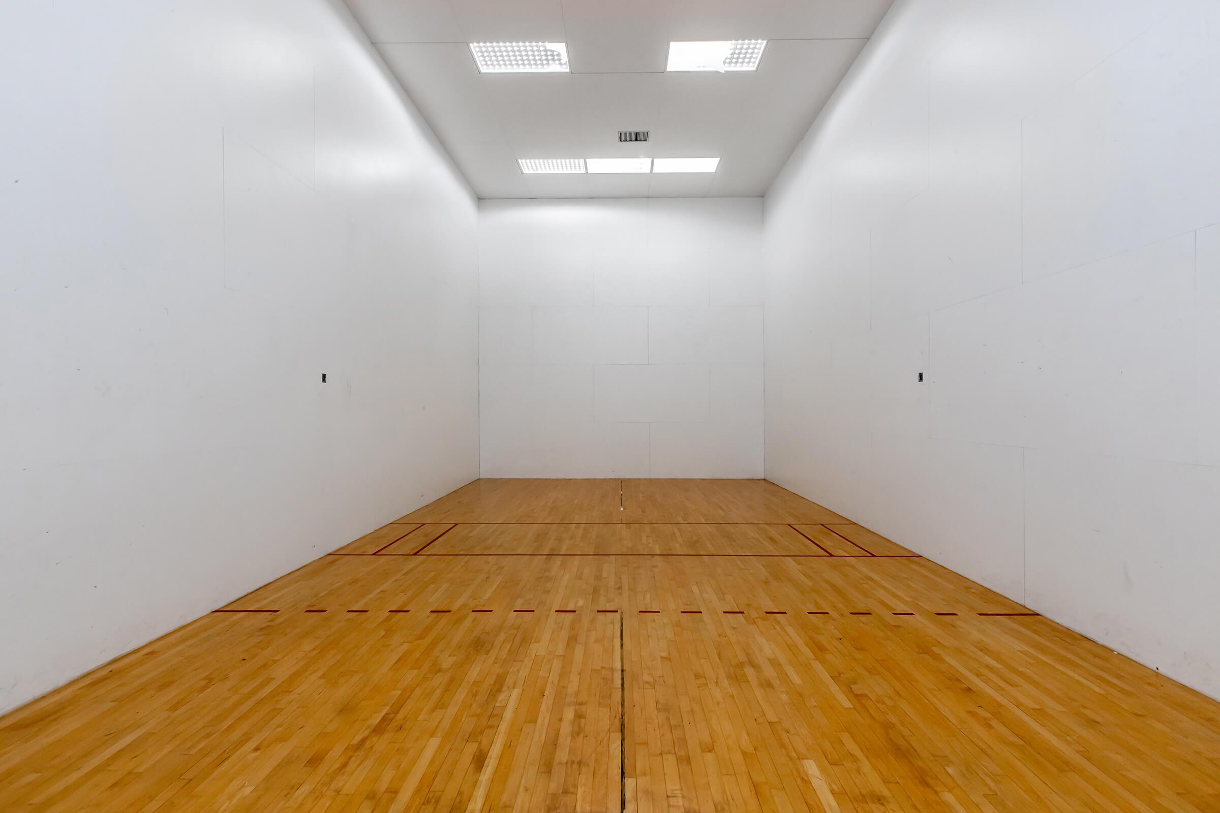 A spacious, empty racquetball court featuring smooth wooden flooring and white walls. The court has marked lines on the floor for gameplay and is brightly lit with overhead lights. There are no obstacles or equipment visible in the space.