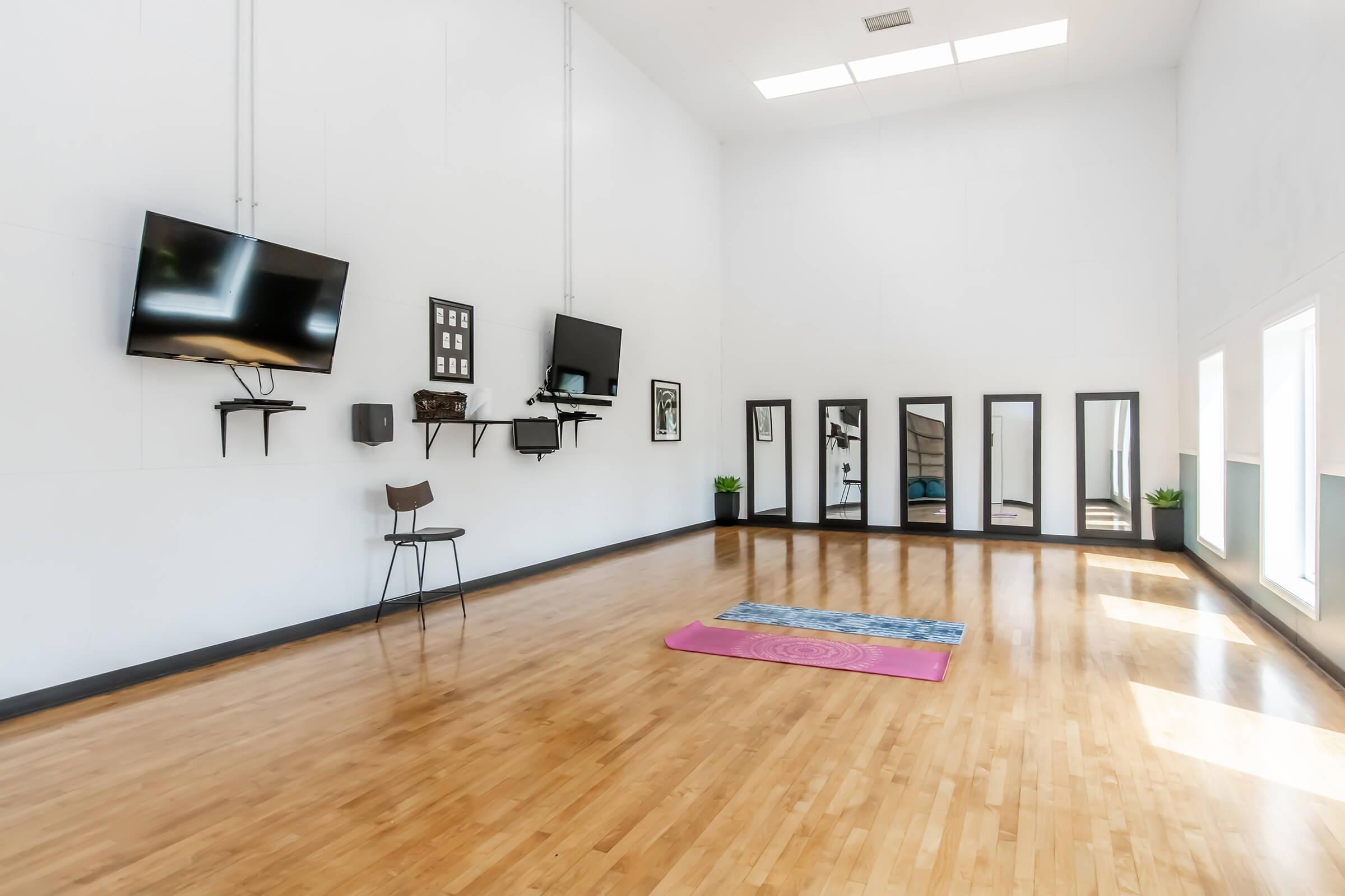 A spacious, well-lit studio featuring wooden flooring, two mounted televisions, and a chair. The walls are adorned with framed pictures and mirrors. A yoga mat is laid out on the floor, and there are small plants in the background, creating a calm and inviting atmosphere.