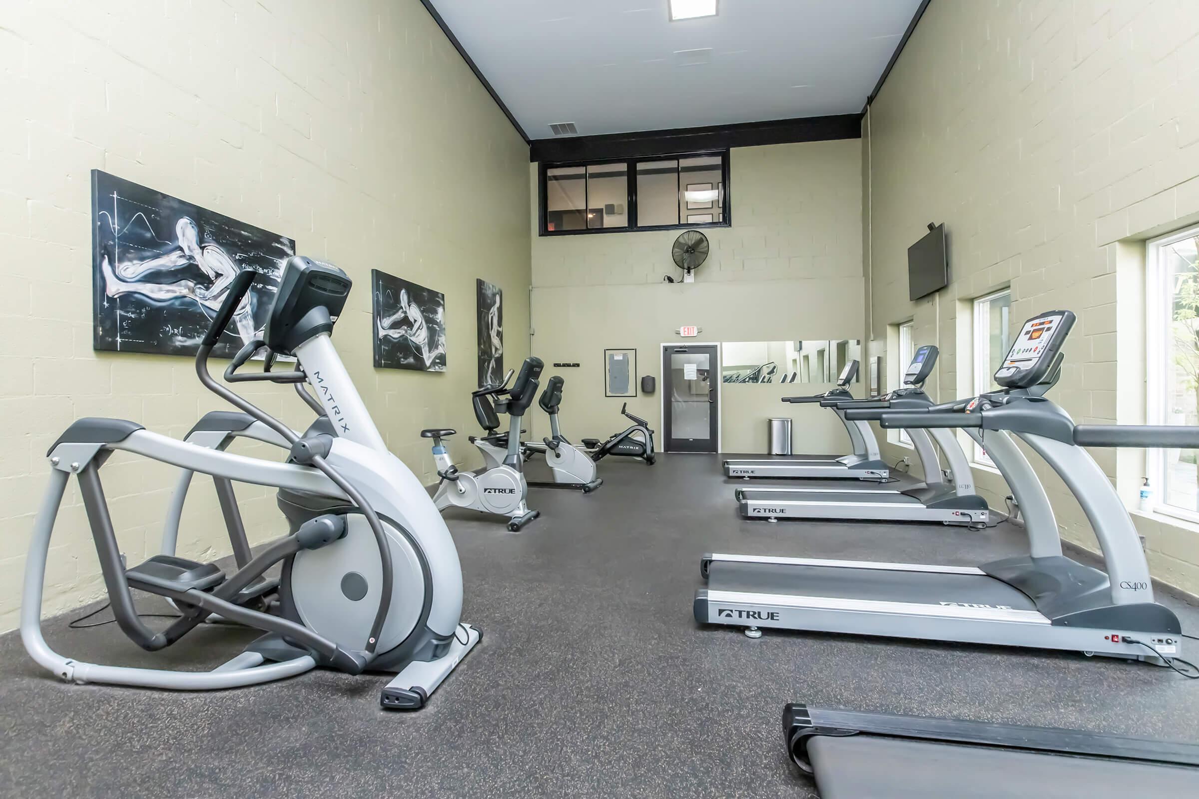 A spacious fitness room featuring multiple exercise machines, including treadmills and ellipticals. Large windows provide natural light, and motivational artwork adorns the walls. The environment is clean and well-maintained, ideal for workouts.