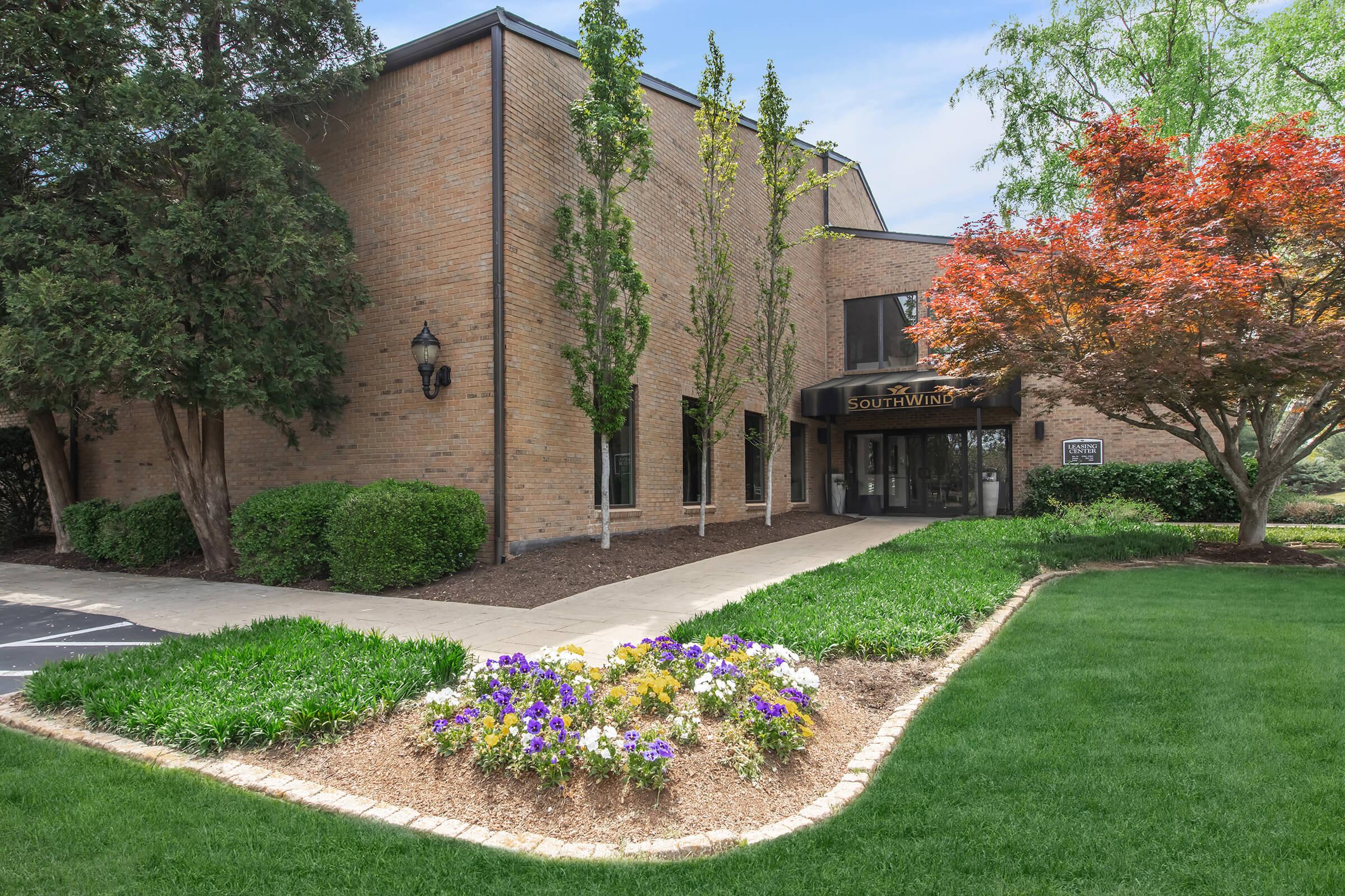 A brick building labeled "Southwind" surrounded by well-maintained landscaping featuring green grass, colorful flower beds, and small trees. The entrance is visible with large windows and a welcoming path.