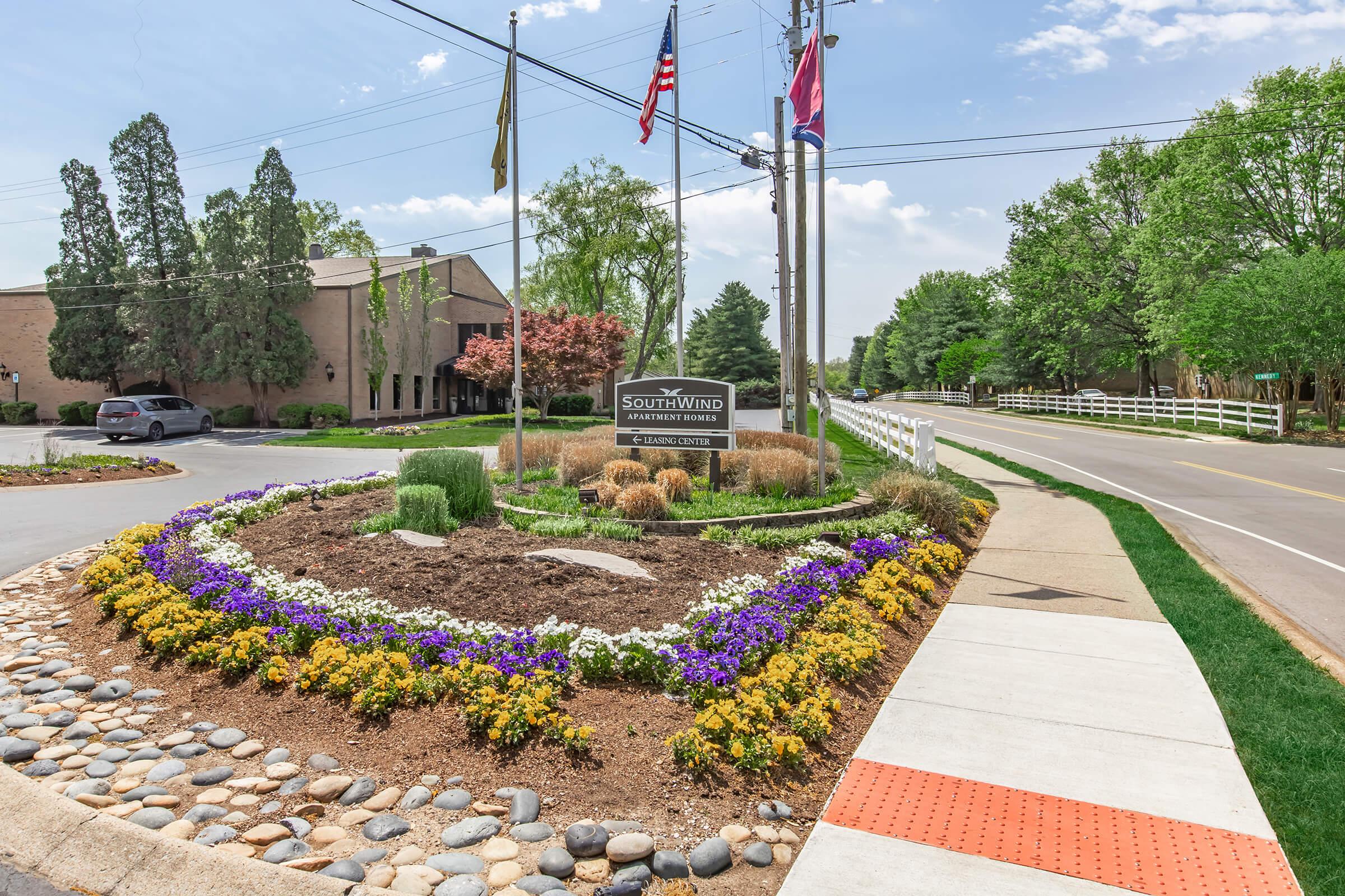 A landscaped roundabout featuring colorful flowers and decorative stones, with a sign for "Southwind," surrounded by green trees and a clear blue sky. American flags are visible on poles nearby, and there is a sidewalk leading to the road.