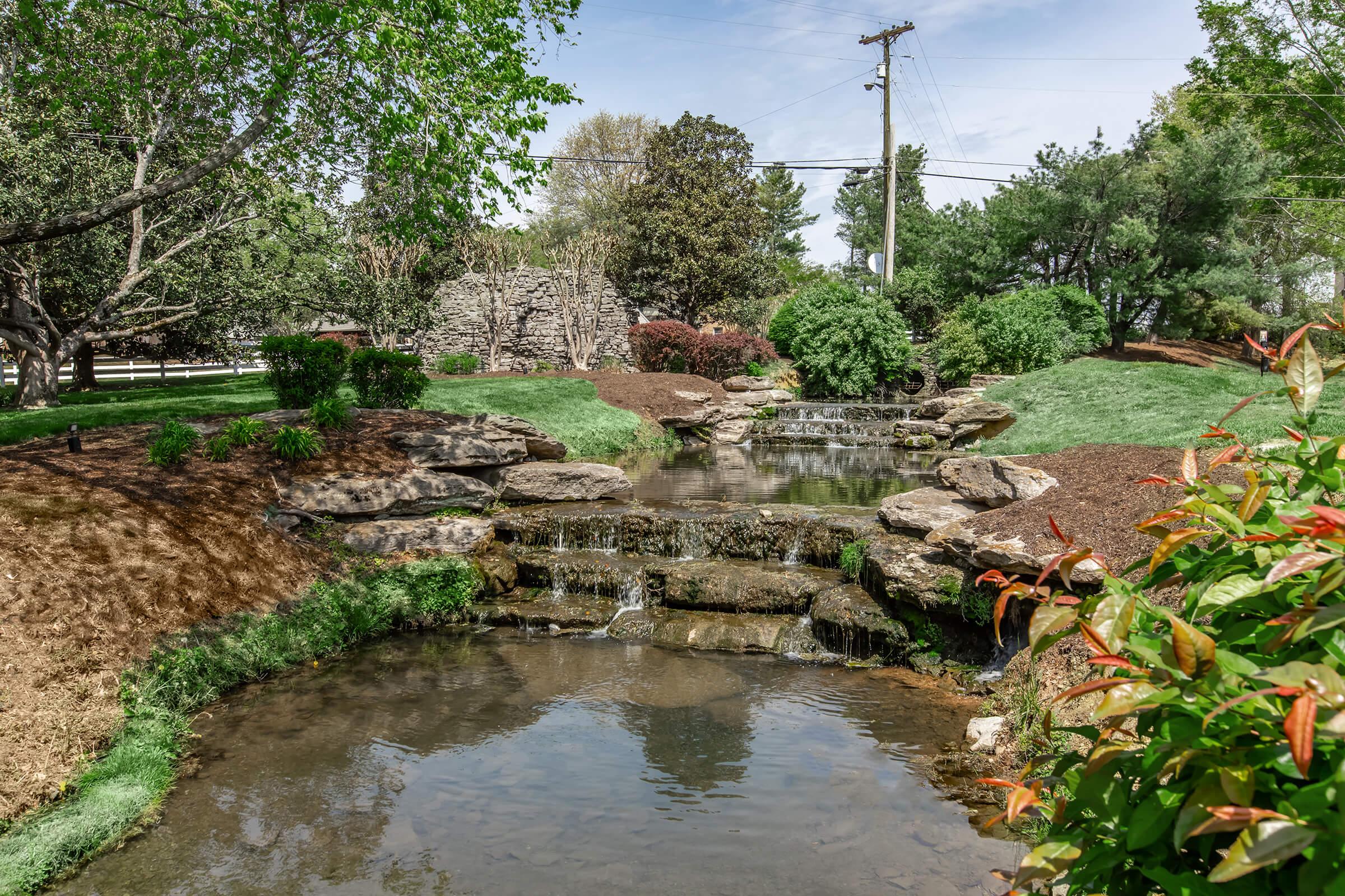 A serene landscape featuring a small waterfall cascading over rocks into a clear pond. Lush greenery surrounds the water, with well-maintained shrubs and trees in the background. The scene is bright and inviting, showcasing a peaceful outdoor setting.