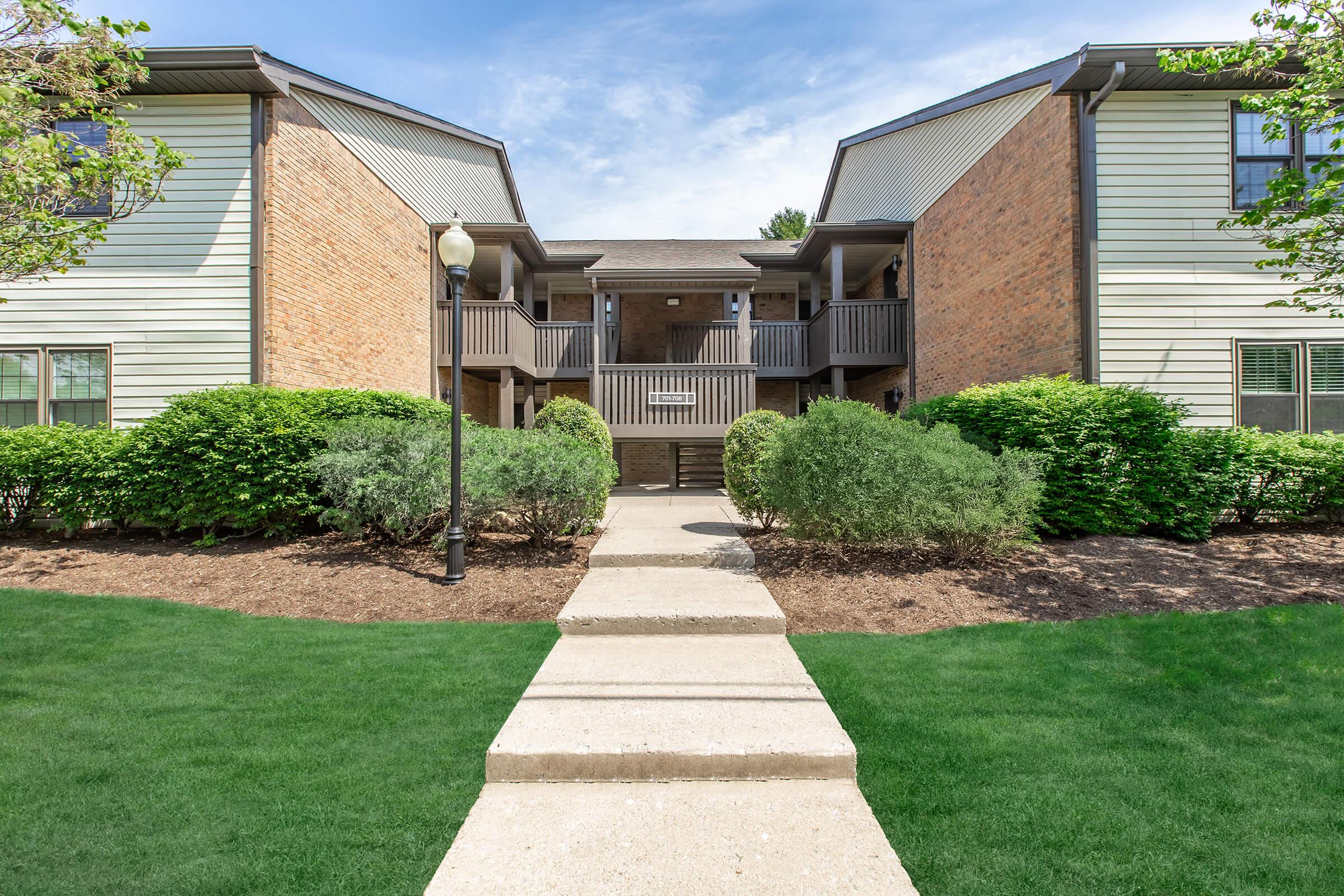 View of a residential apartment complex featuring two buildings with a walkway leading to the entrance. The area is landscaped with greenery and shrubbery, under a clear blue sky. The architecture includes a mix of brick and siding materials, with balconies visible on the upper floors.