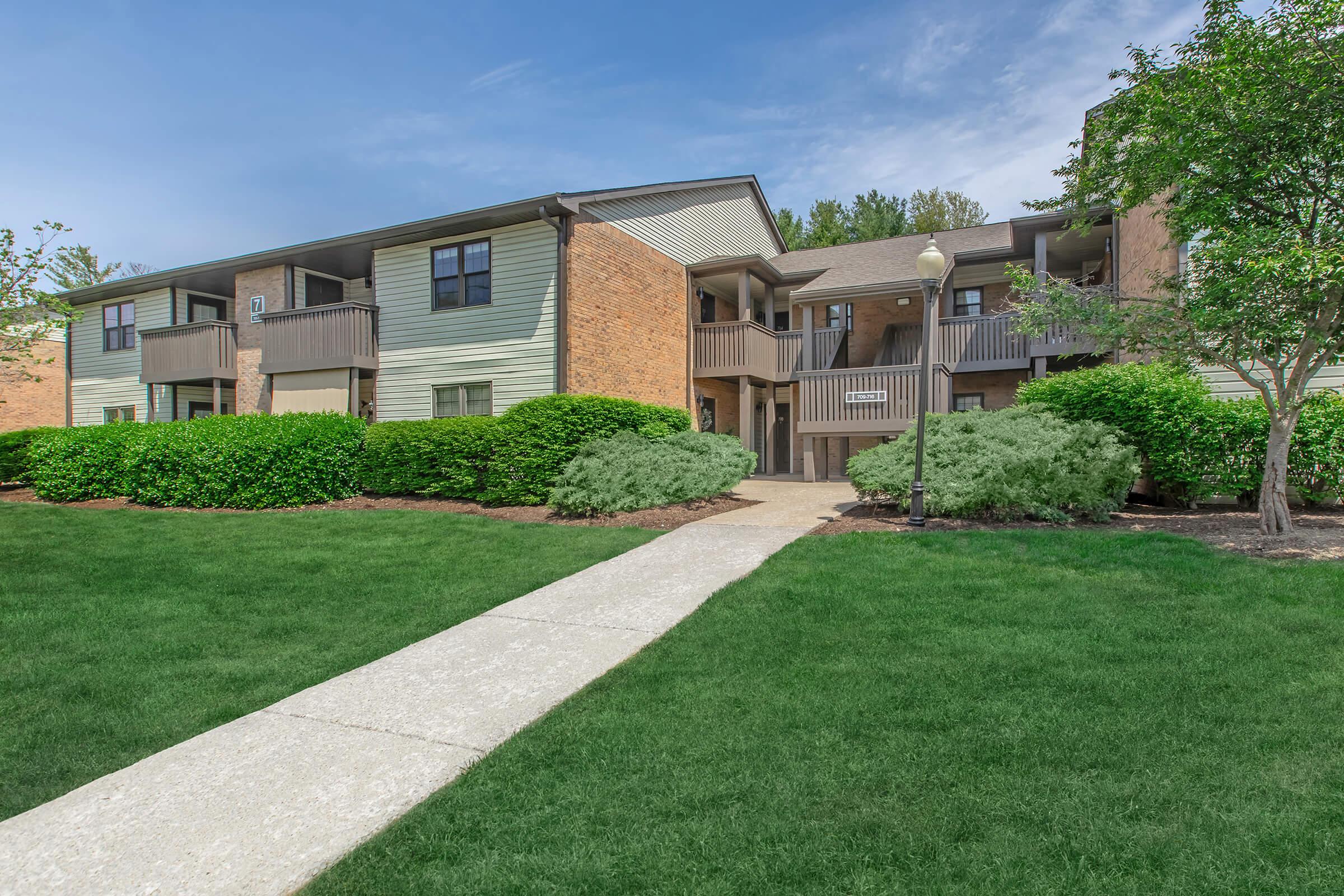 A well-maintained apartment complex with two-level buildings. Green lawns and shrubs surround the buildings, with a concrete walkway leading to the entrance. Clear blue skies above add to the inviting atmosphere.