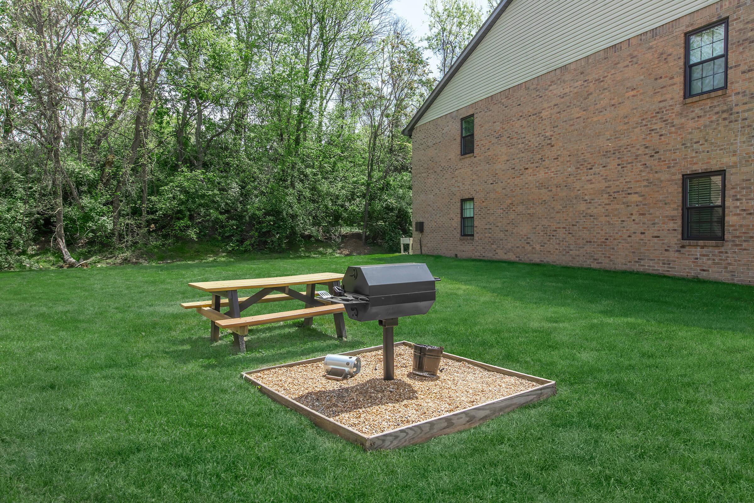 A grassy backyard featuring a picnic table and a charcoal grill on a gravel base, positioned near a brick building. Lush trees and greenery create a natural backdrop, making it an inviting outdoor space for gatherings and barbecues.