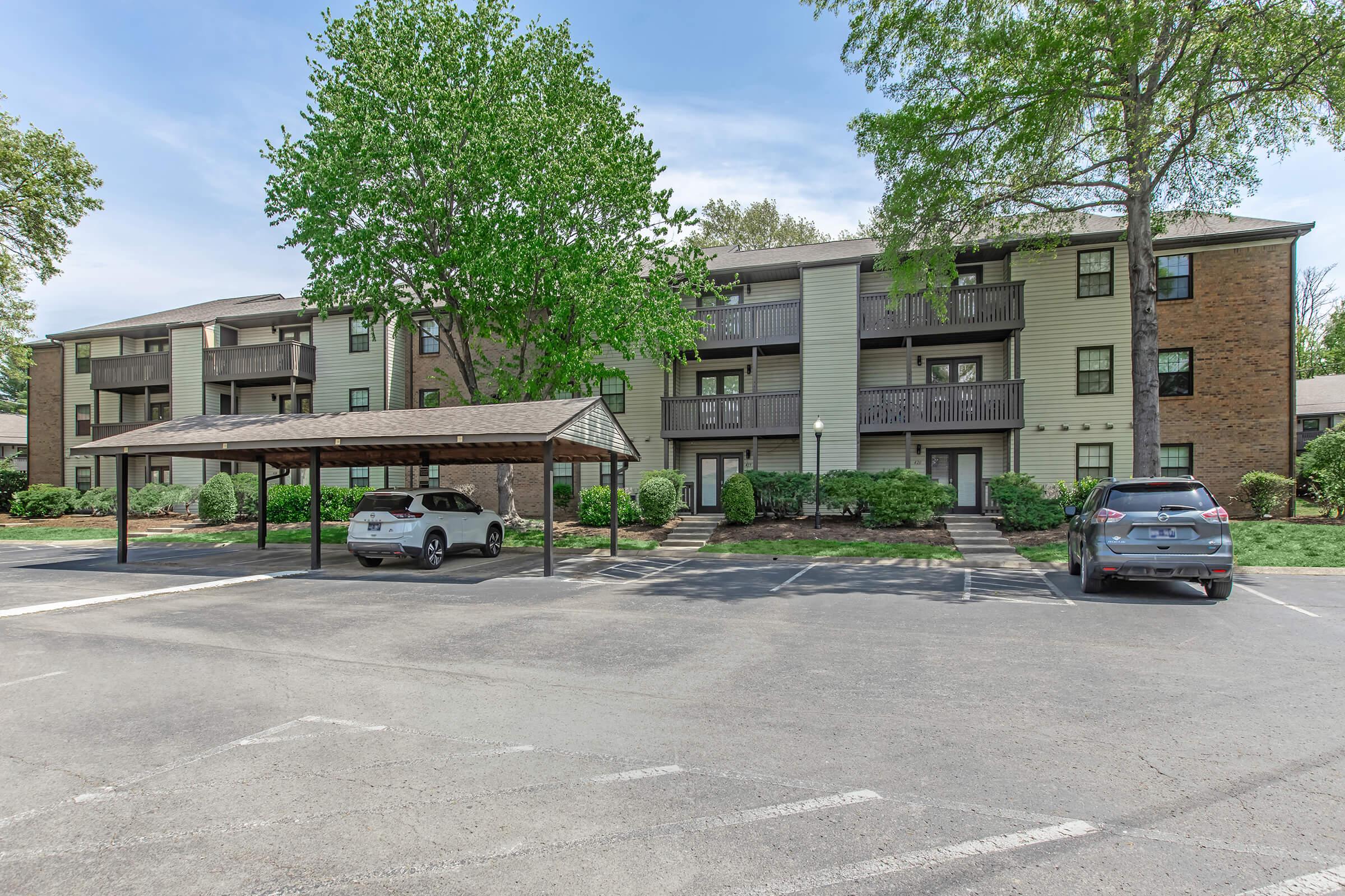 A multi-story apartment building with a light green exterior, surrounded by trees and shrubs. There are two parked cars in the foreground, one under a carport. The area is paved with marked parking spaces, and the sky is clear and blue.