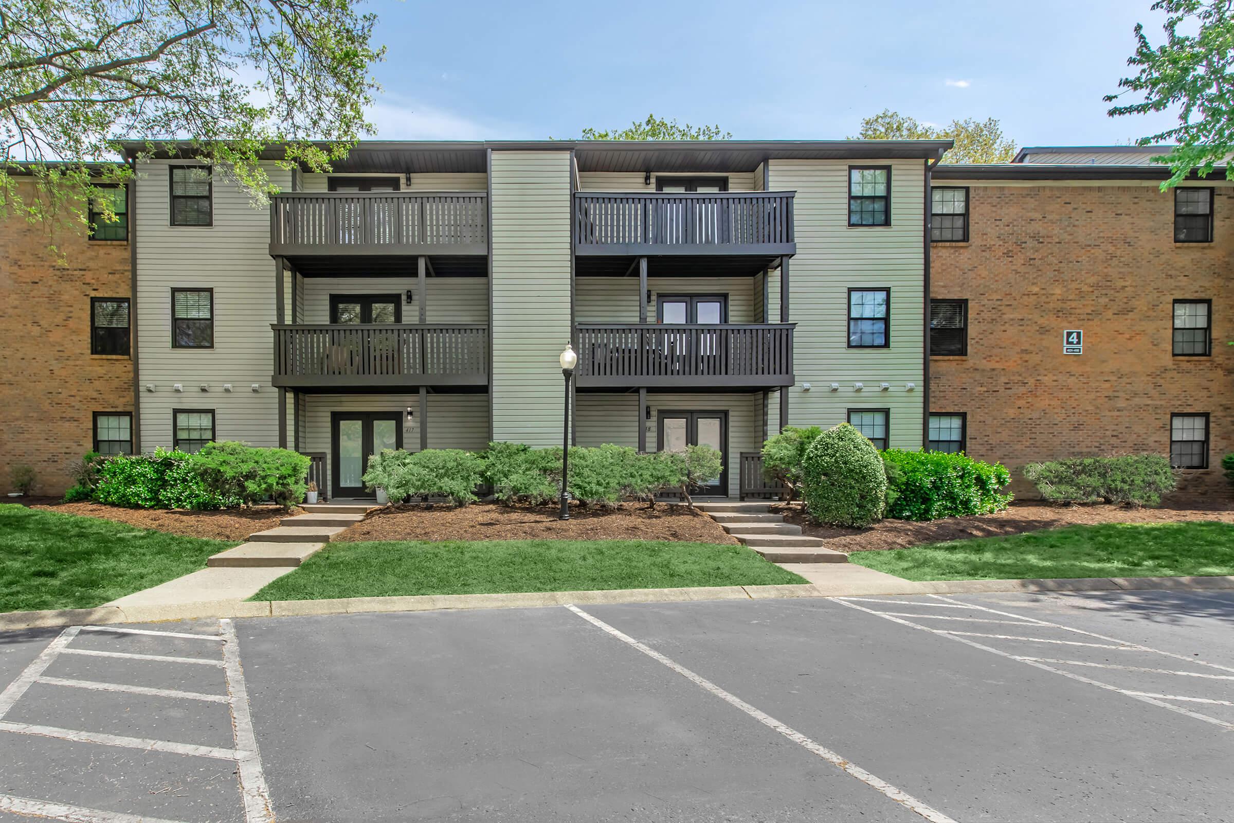 A two-story apartment building featuring a brick exterior with gray siding. The building has multiple balconies and is surrounded by landscaped greenery, including shrubs and trees. A parking area is visible in the foreground, with designated parking spaces. The sky is clear and blue.