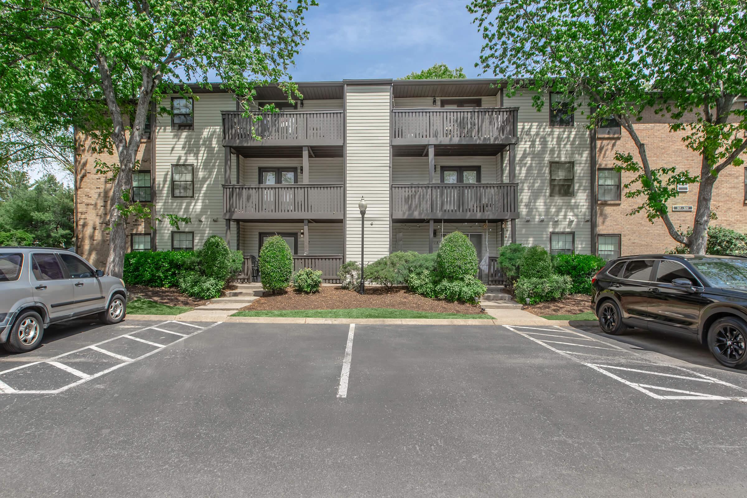A two-story apartment building with balconies, surrounded by greenery. Two parking spaces are visible in the foreground, with a gray SUV and a black SUV parked. The building has light-colored siding and dark wooden railings, with trees providing shade on either side.