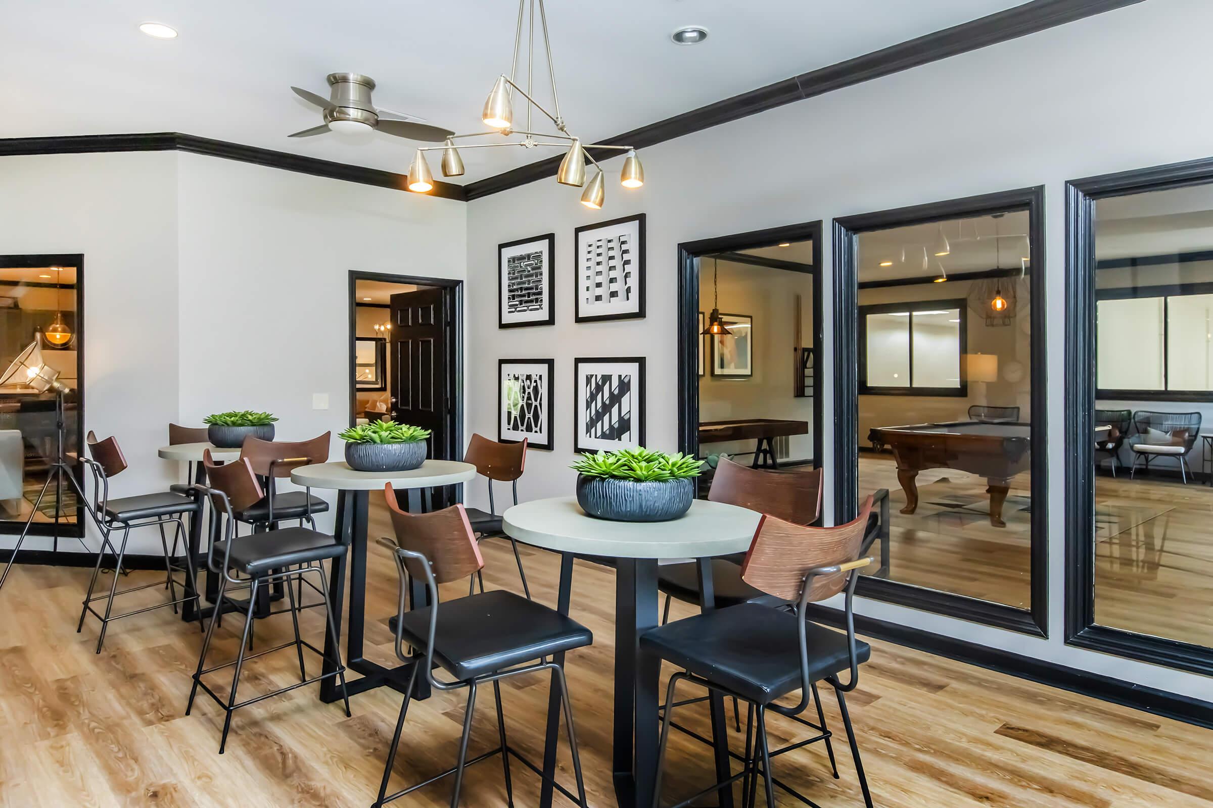 Modern lounge area with several high-top tables and stylish chairs. The walls feature framed artwork and mirrors, creating a spacious feel. Plants adorn the tables, and a ceiling fan provides a contemporary touch. The flooring is wooden, enhancing the inviting atmosphere.