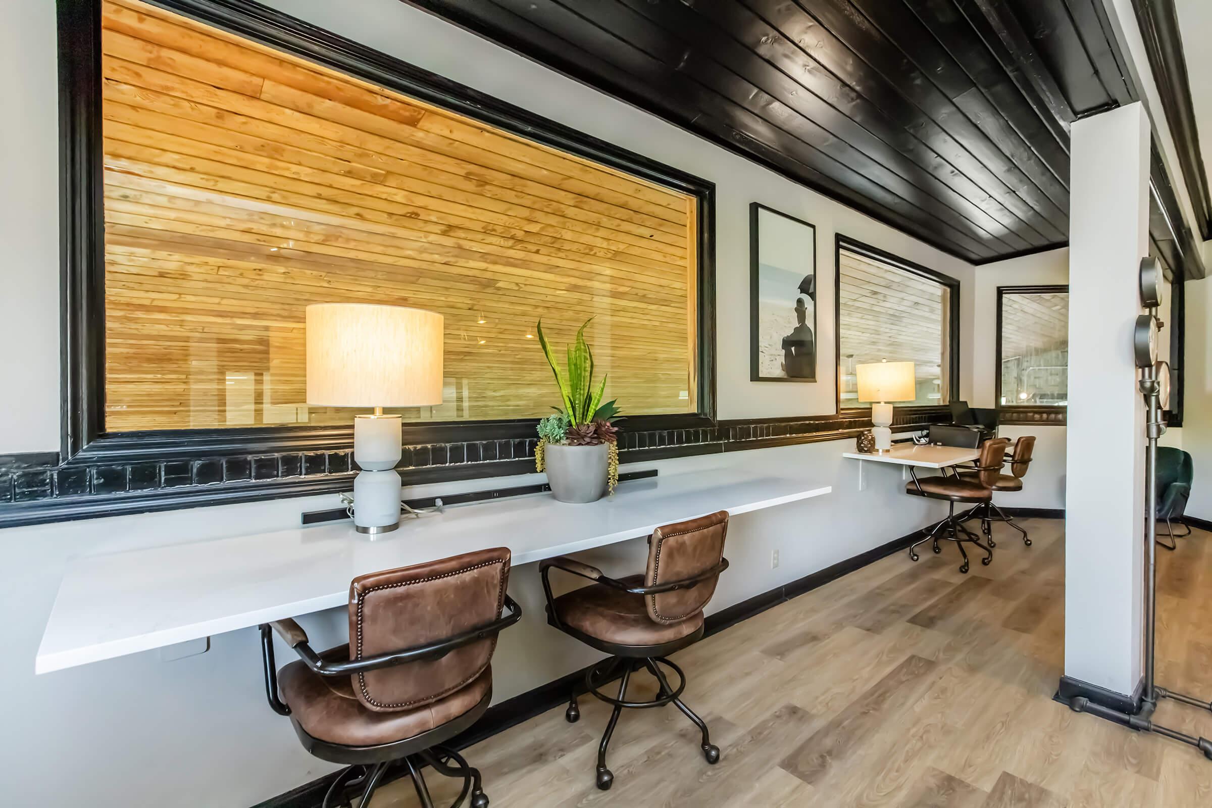 Modern workspace featuring two brown leather chairs at a white long table, illuminated by a stylish lamp. Large windows with wooden paneling in the background provide natural light. Minimalist decor adds a contemporary touch to the office environment.