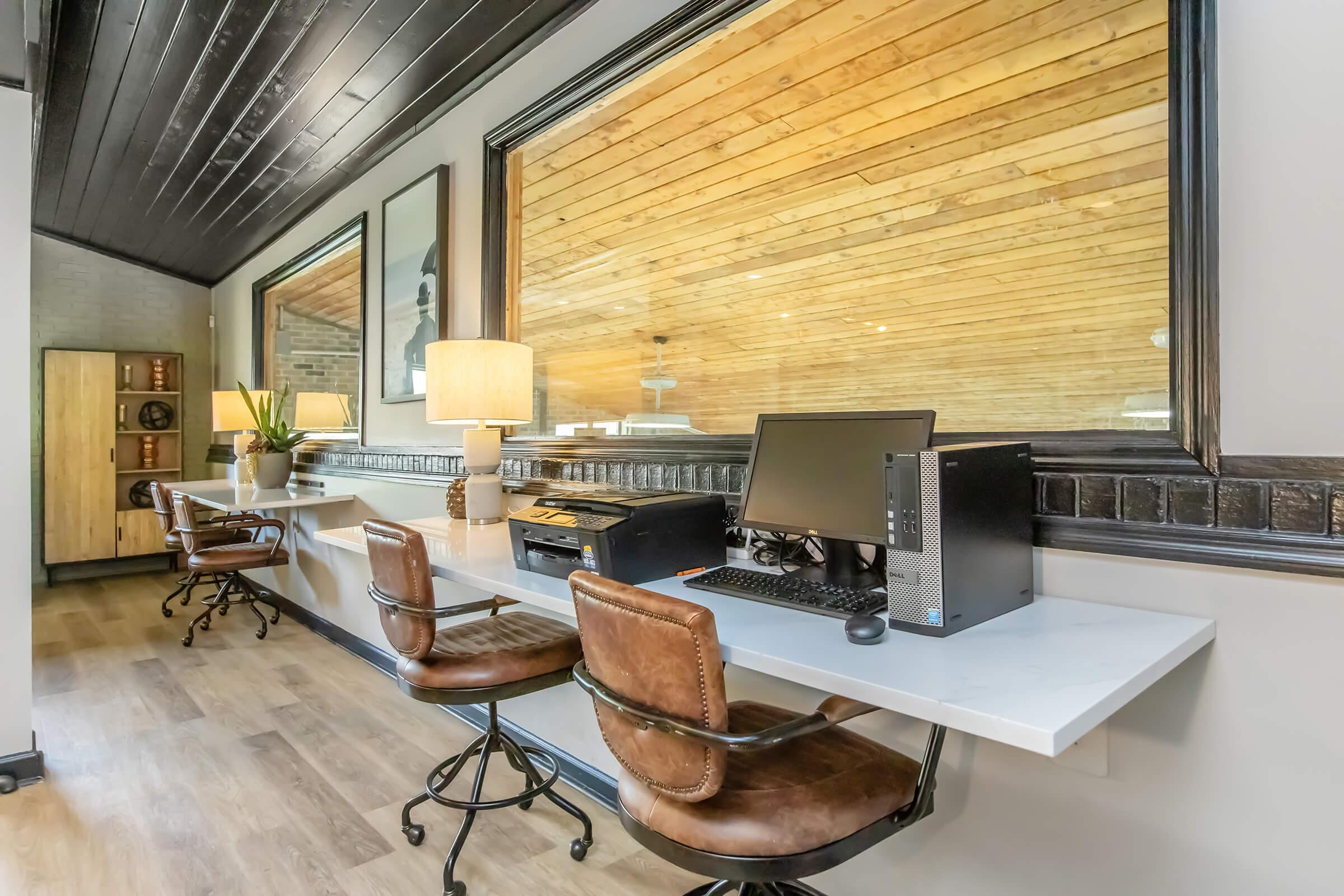 Modern workspace featuring two desks with brown leather chairs, a desktop computer, and a printer. Large windows with wooden accents allow natural light to flood the area, enhancing the warm, inviting atmosphere. The room has light-colored walls and wooden flooring, creating a stylish and functional environment.
