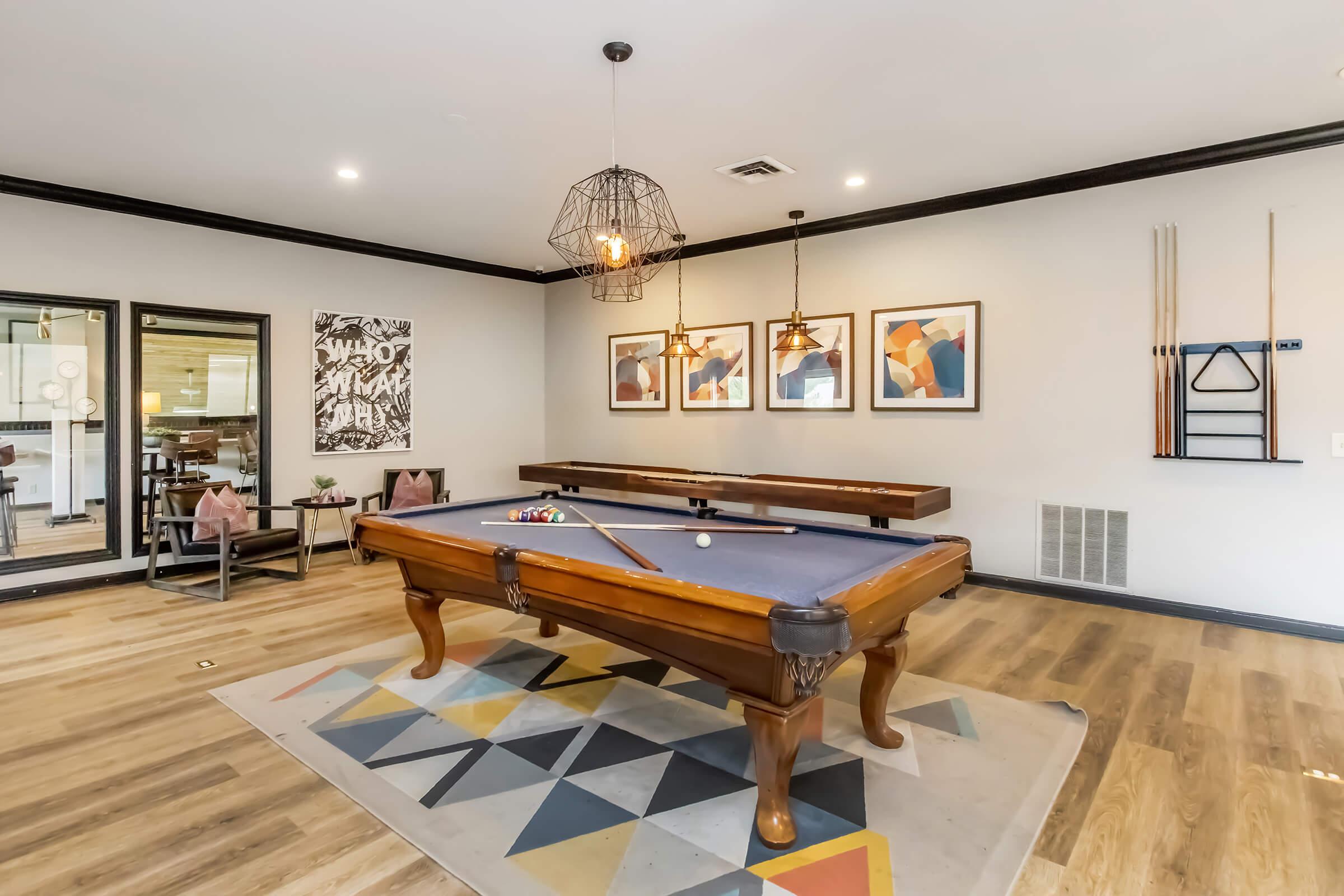 A cozy game room featuring a large pool table with blue felt, surrounded by modern artwork on the walls. The space includes stylish lighting fixtures, wooden flooring, and a geometric patterned rug, with additional seating areas in the background.