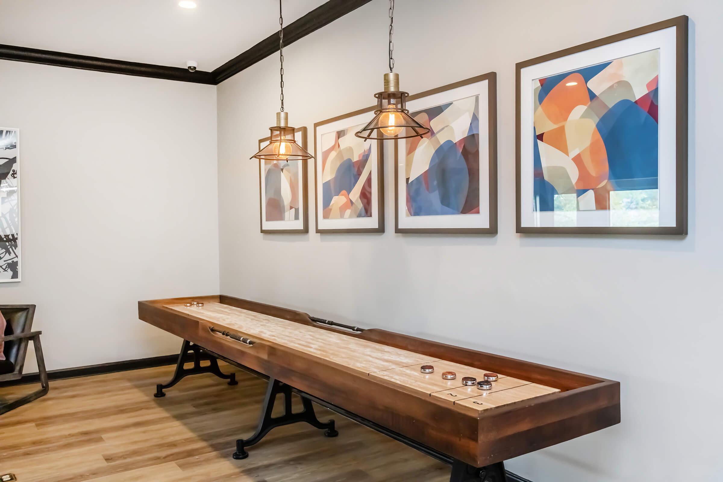 A stylish interior featuring a long wooden shuffleboard table with several pucks on it. The background includes three framed abstract artworks. The space is adorned with modern pendant lights and has a warm wooden floor, creating a cozy atmosphere.
