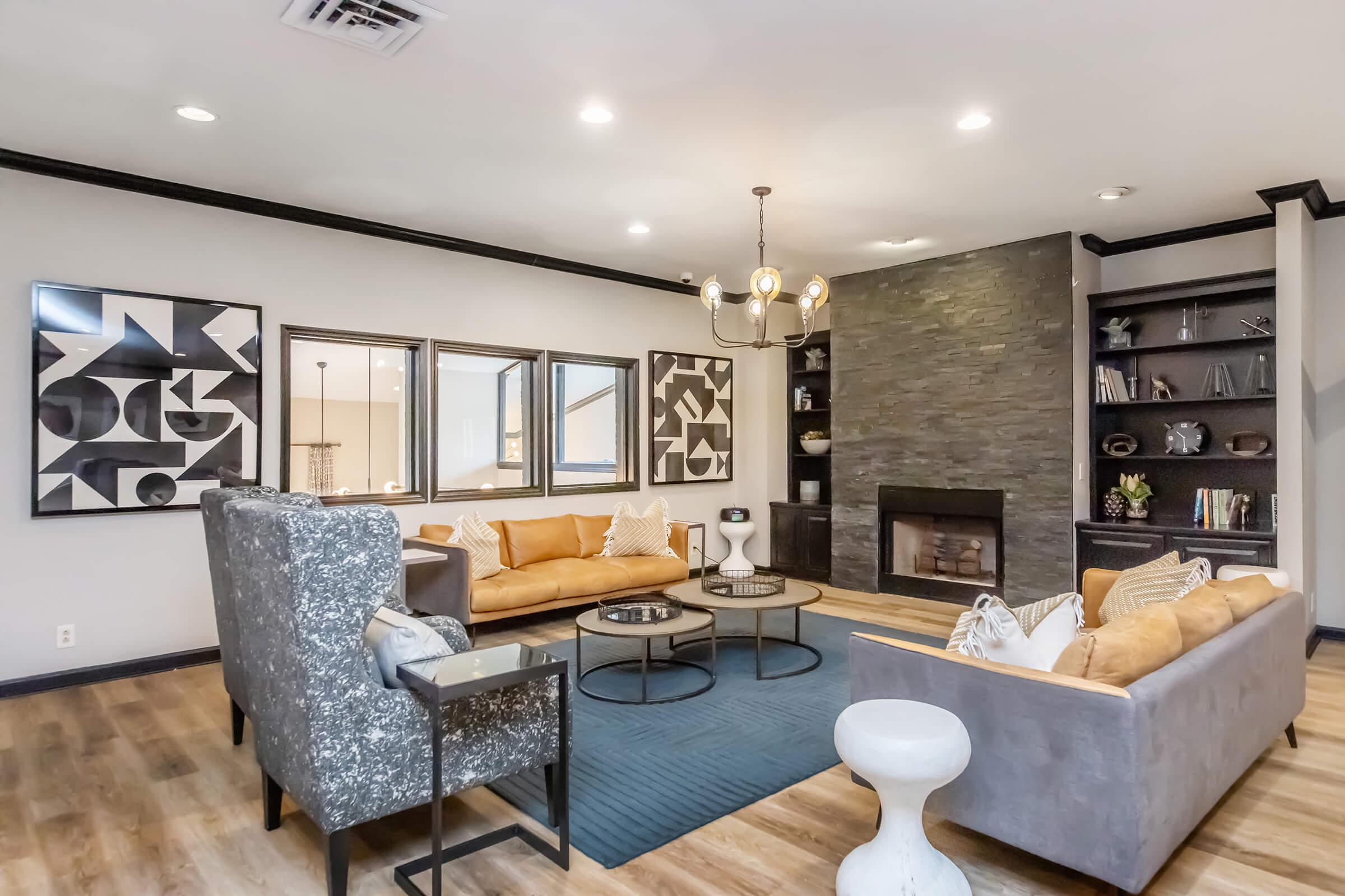 A cozy living room featuring a yellow leather sofa and a gray couch, complemented by patterned throw pillows. A modern chandelier hangs above, and a stone fireplace with a dark mantel is visible. Decorative mirrors and abstract artwork adorn the walls, and a blue area rug adds a stylish touch to the wooden floor.
