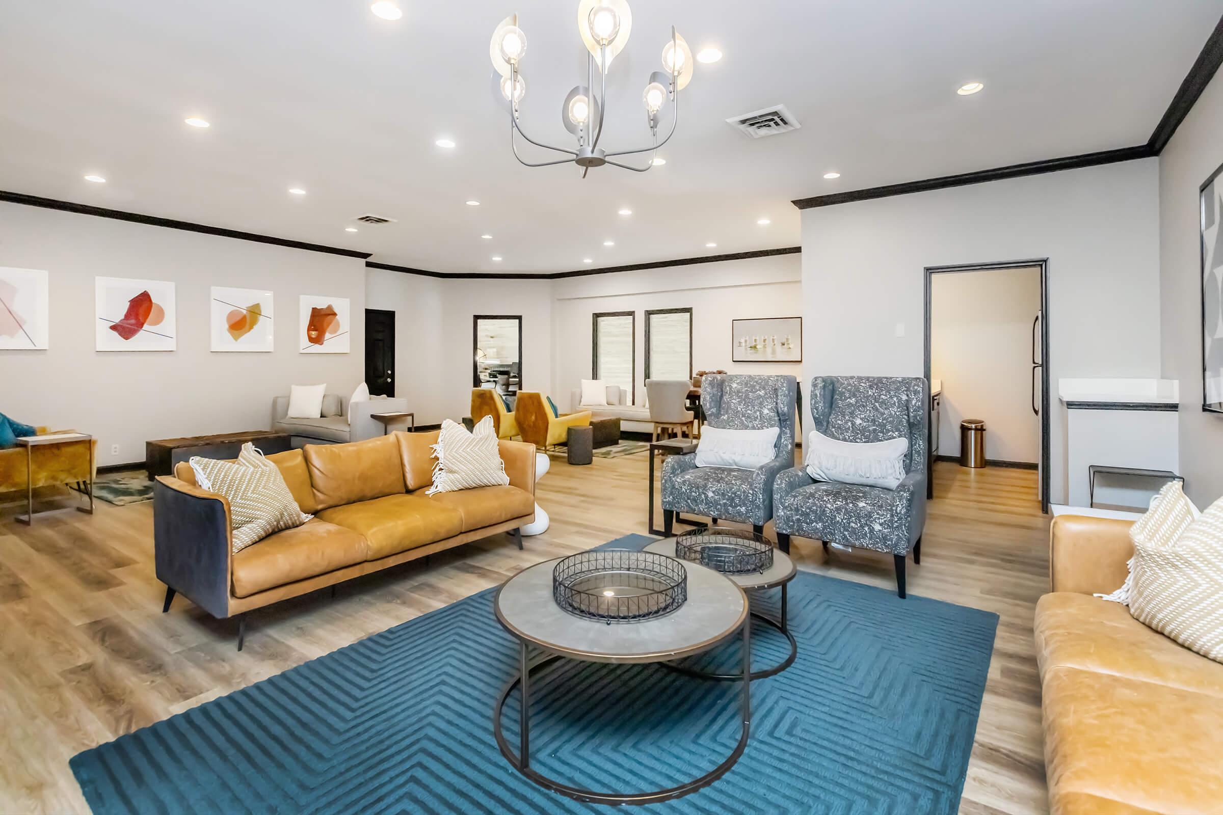 A modern lounge area featuring a mix of furniture styles. There are leather sofas in warm tones, patterned armchairs, and a round coffee table on a blue textured rug. Soft lighting from overhead fixtures illuminates art on the walls and creates an inviting atmosphere.