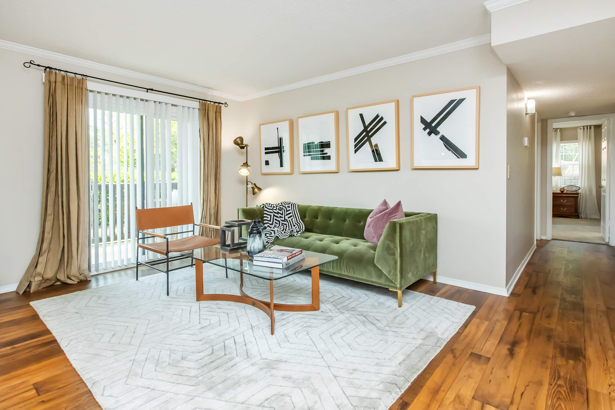 A stylish living room featuring a green velvet sofa with decorative pillows, a modern chair, and a geometric rug. The walls display framed abstract artwork, and there's natural light flowing through sheer curtains. The hardwood floor adds warmth, while a side table holds books and decor.