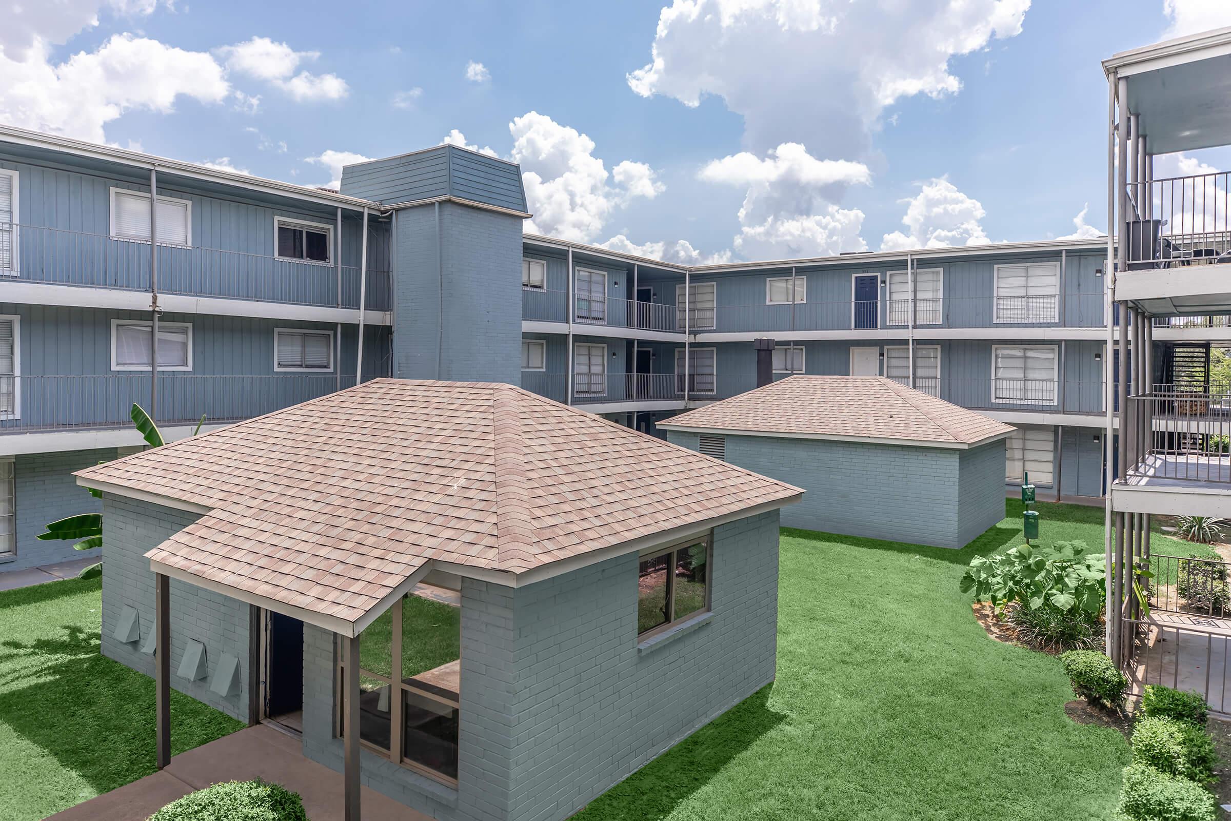 A view of a residential complex featuring two single-story buildings with brown shingled roofs surrounded by green grass. The buildings are painted in light blue, and there's a larger multi-story building in the background. The sky is partly cloudy, creating a bright atmosphere.