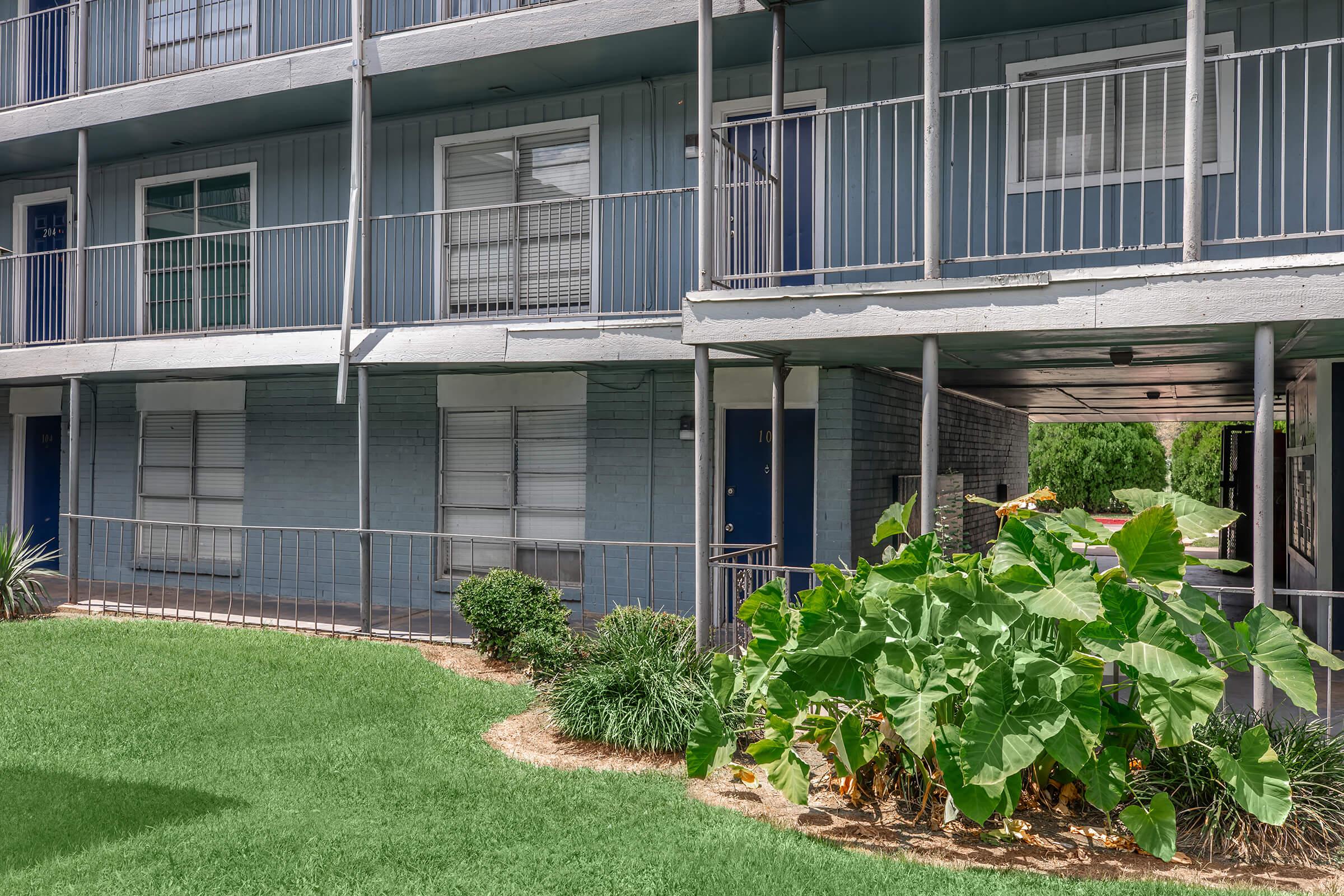 Ground-level view of a residential building with blue siding and multiple balconies. The area features a well-maintained garden with lush greenery and large plants. The setting is tidy, with grass and shrubs nearby, creating a welcoming atmosphere.