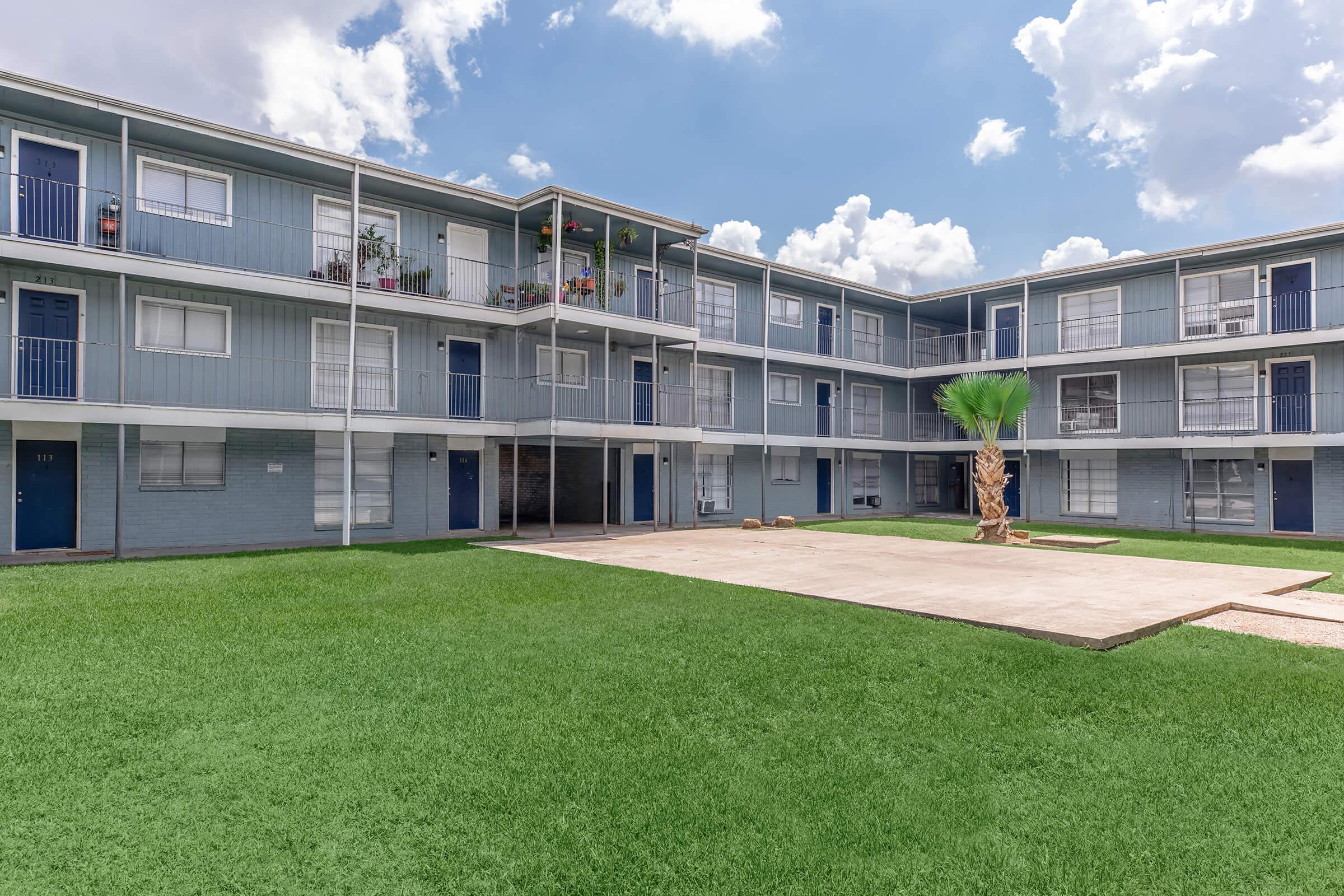 A view of a multi-story apartment building with blue siding and balconies, surrounded by green grass. The courtyard features a small paved area and a palm tree. The sky is partly cloudy, adding a bright atmosphere to the scene.