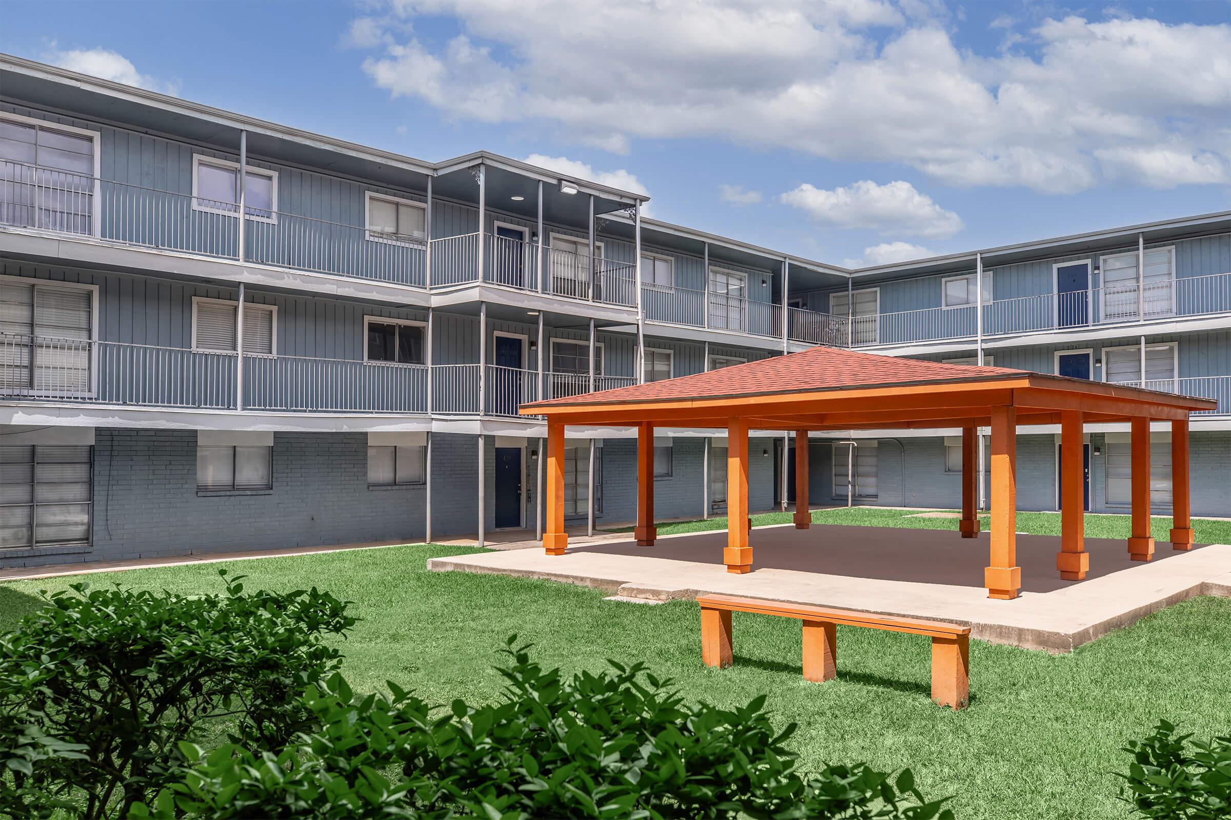 A brightly colored gazebo with an orange roof and columns is situated in the center of a grassy courtyard surrounded by two-story blue apartment buildings. The sky above is partly cloudy, creating a serene outdoor space for residents.