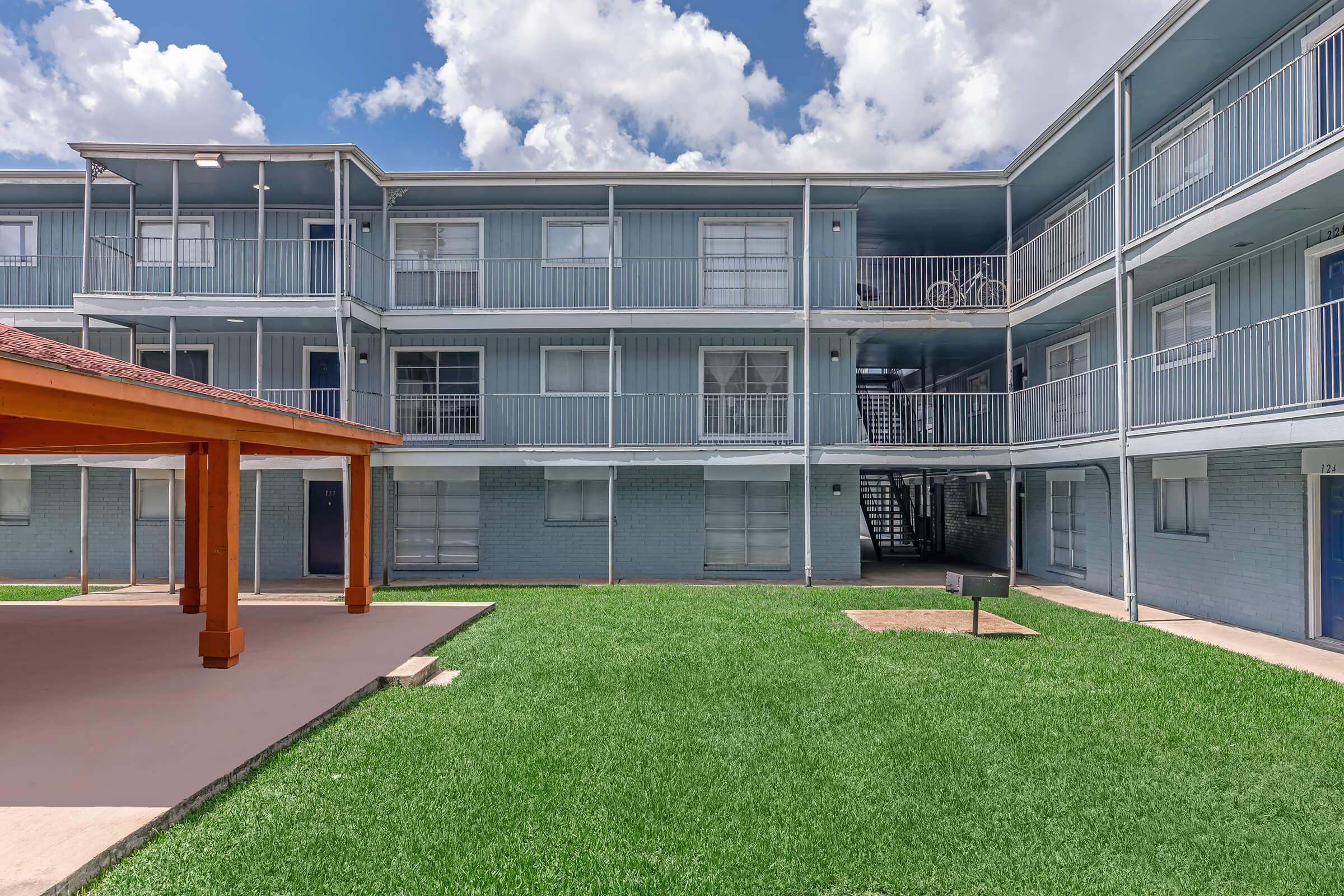 A multi-story apartment building with blue exterior walls, featuring balconies and large windows. The courtyard in front has well-maintained green grass, a grill area, and a wooden structure for seating. Bright clouds are visible in the sky above.