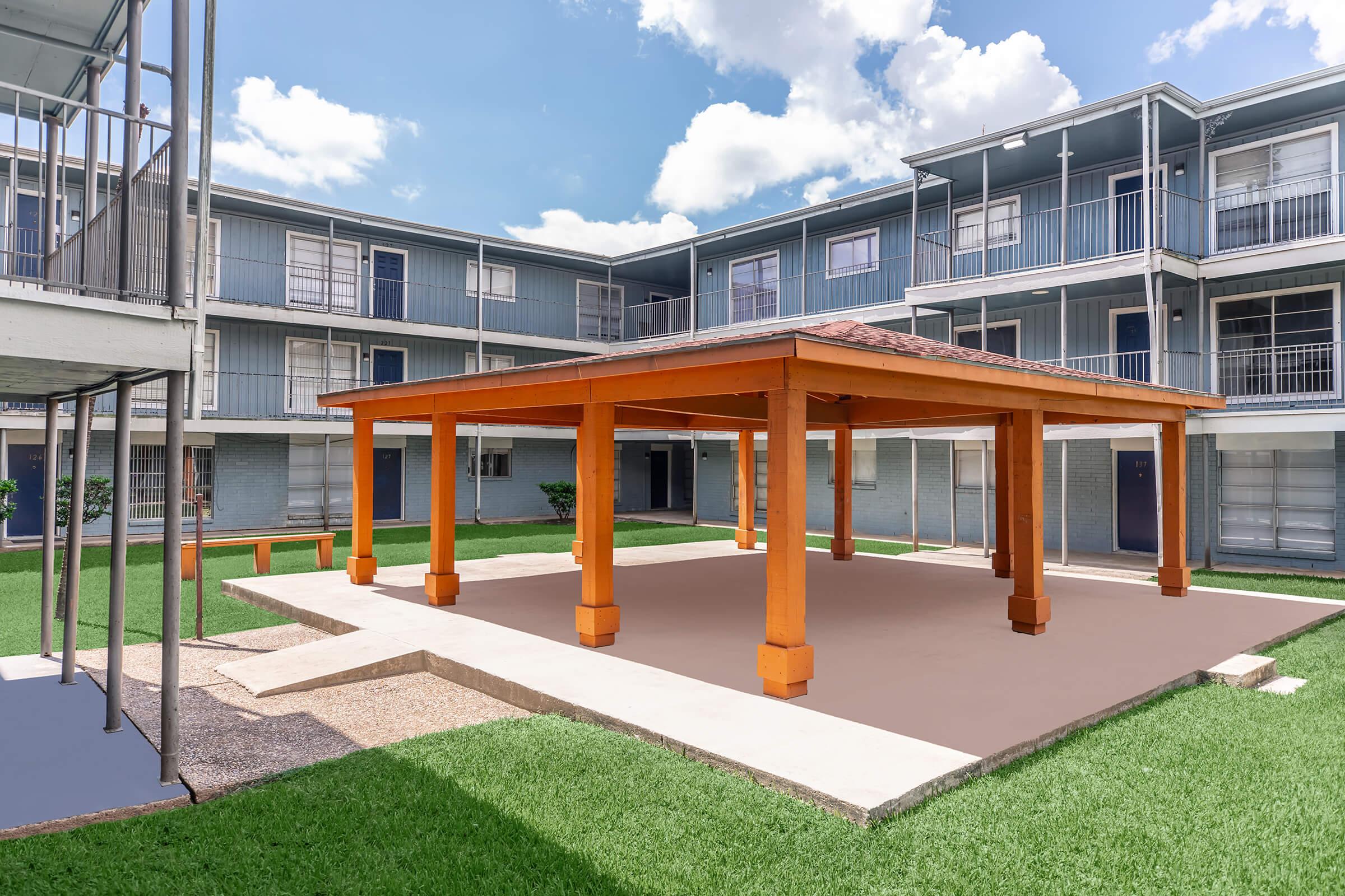A spacious outdoor courtyard with a wooden pavilion featuring orange columns, surrounded by green grass. The area is flanked by two-story buildings, each with multiple balconies. The sky is blue with fluffy white clouds, creating a bright and inviting atmosphere.