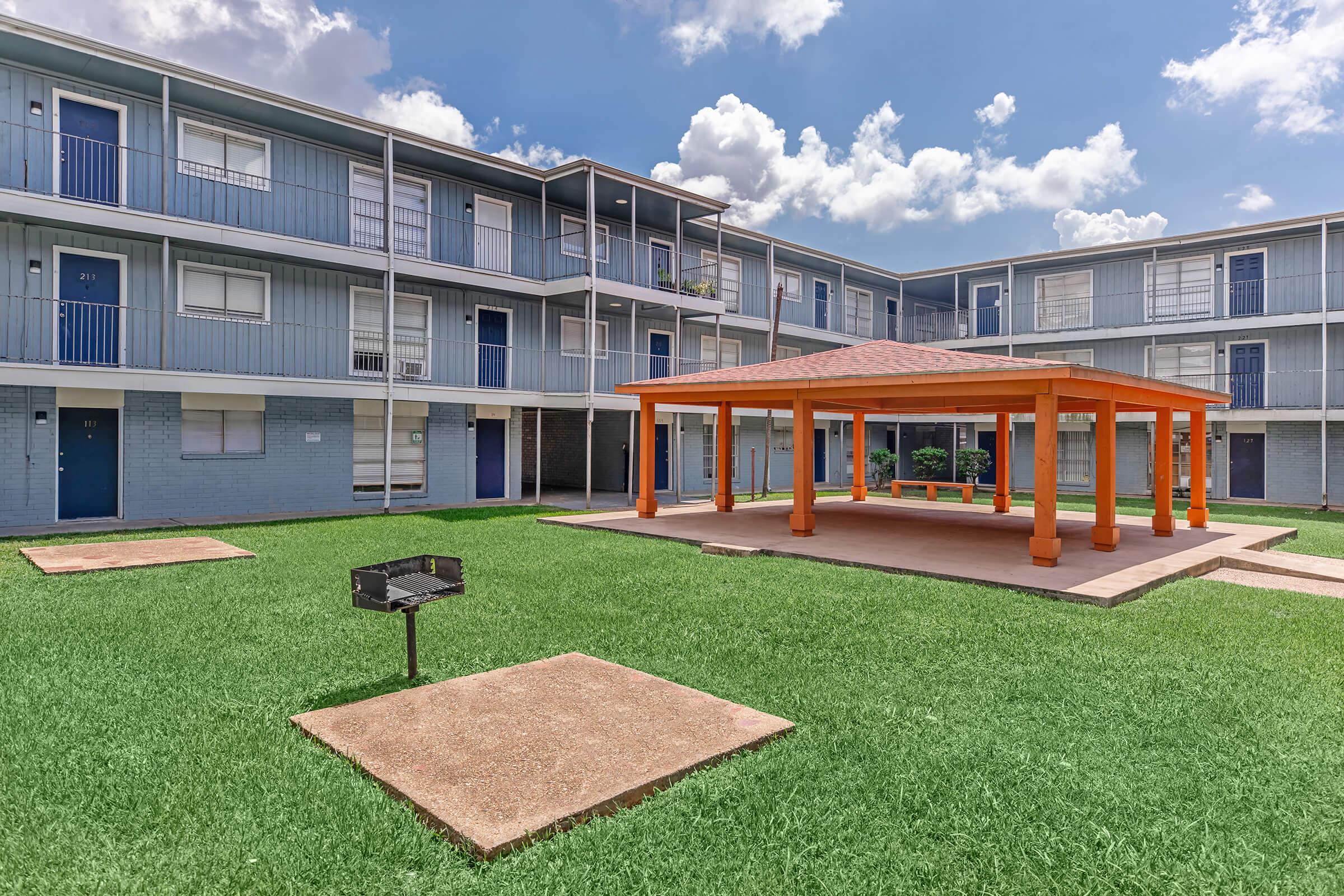 A vibrant outdoor area featuring an orange gazebo surrounded by green grass and square barbecue pits. In the background, there are blue apartment buildings with white railings and balconies, under a partly cloudy sky.