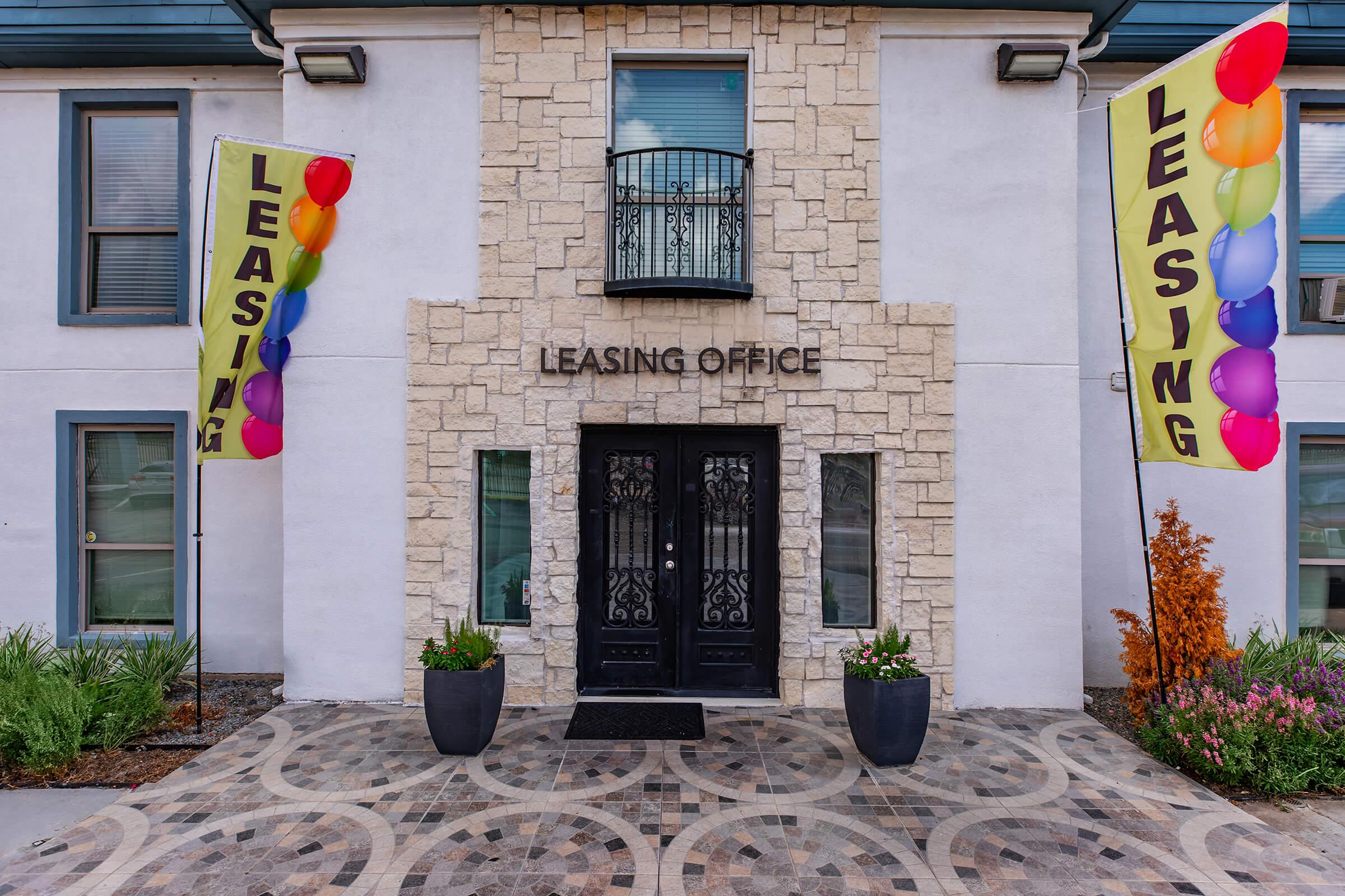 Two large banners with the word "LEASING" in bold yellow are positioned on either side of a modern leasing office entrance. The exterior features a stone facade, double black doors with ornate designs, and a decorative tiled pathway. Colorful balloons add a festive touch to the entrance.