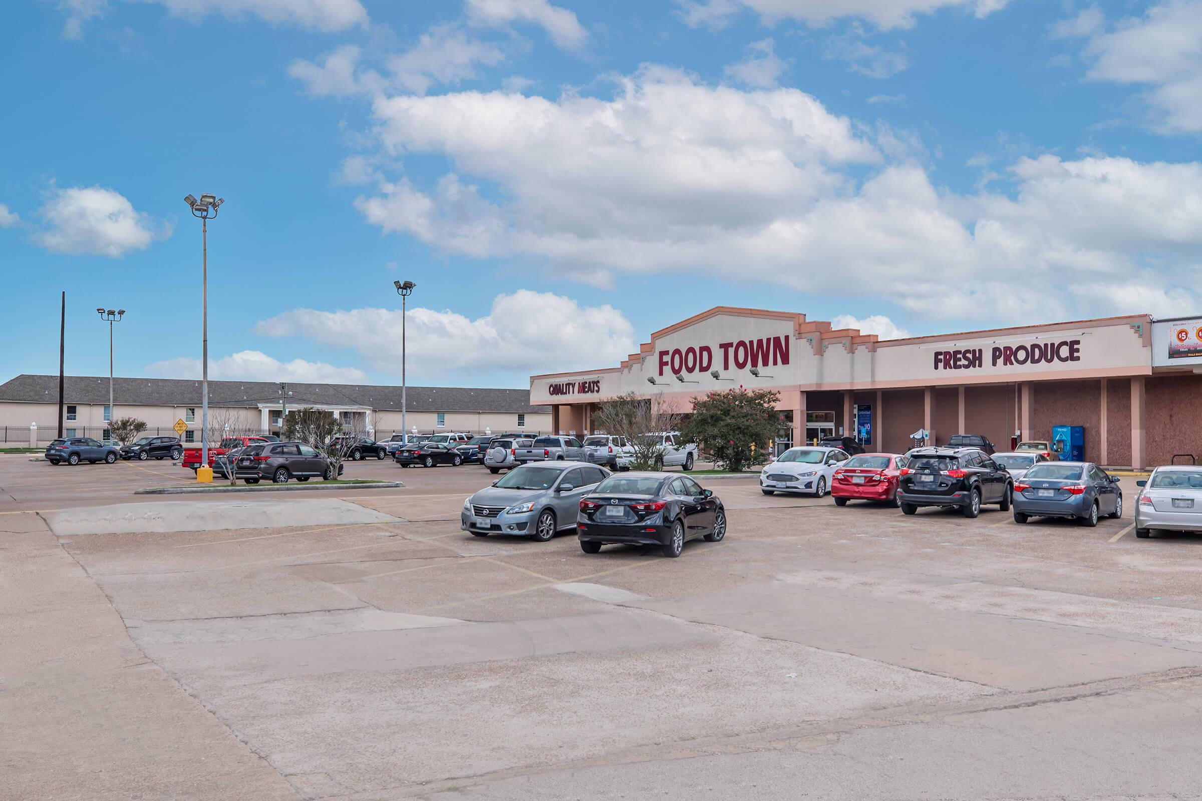Exterior view of a Food Town grocery store featuring a large sign stating "Quality Meat" and "Fresh Produce," with a parking lot filled with various cars. The sky is partly cloudy, and there are light poles in the background.