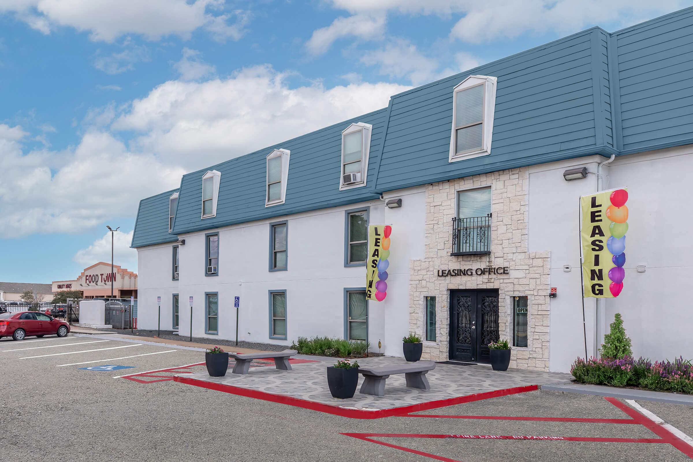 A modern leasing office with a stone entrance, surrounded by colorful balloons. The building features a blue sloped roof and white walls. Parking spaces are visible in front, and there are planters with greenery. Signs indicating "Leasing" hang on either side of the entrance.
