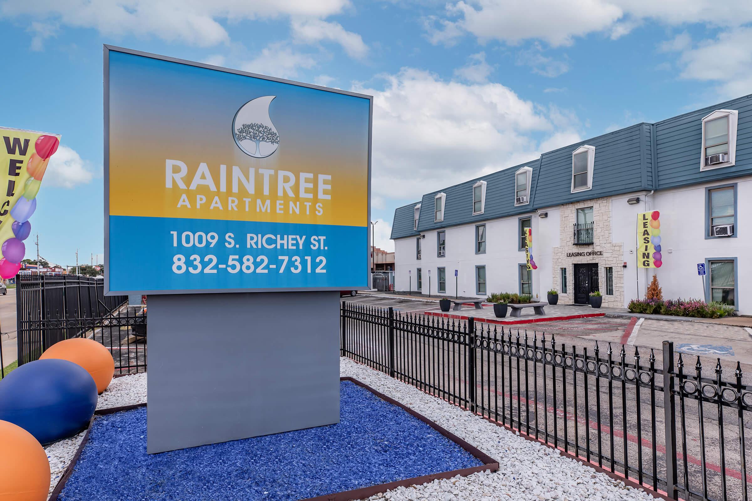 Sign for Raintree Apartments located at 1009 S. Richey St. with a phone number 832-582-7312. The sign features a wave design and is placed in front of a two-story building with a leasing office. Colorful balloons are visible in the foreground, and the area has a well-maintained landscape.