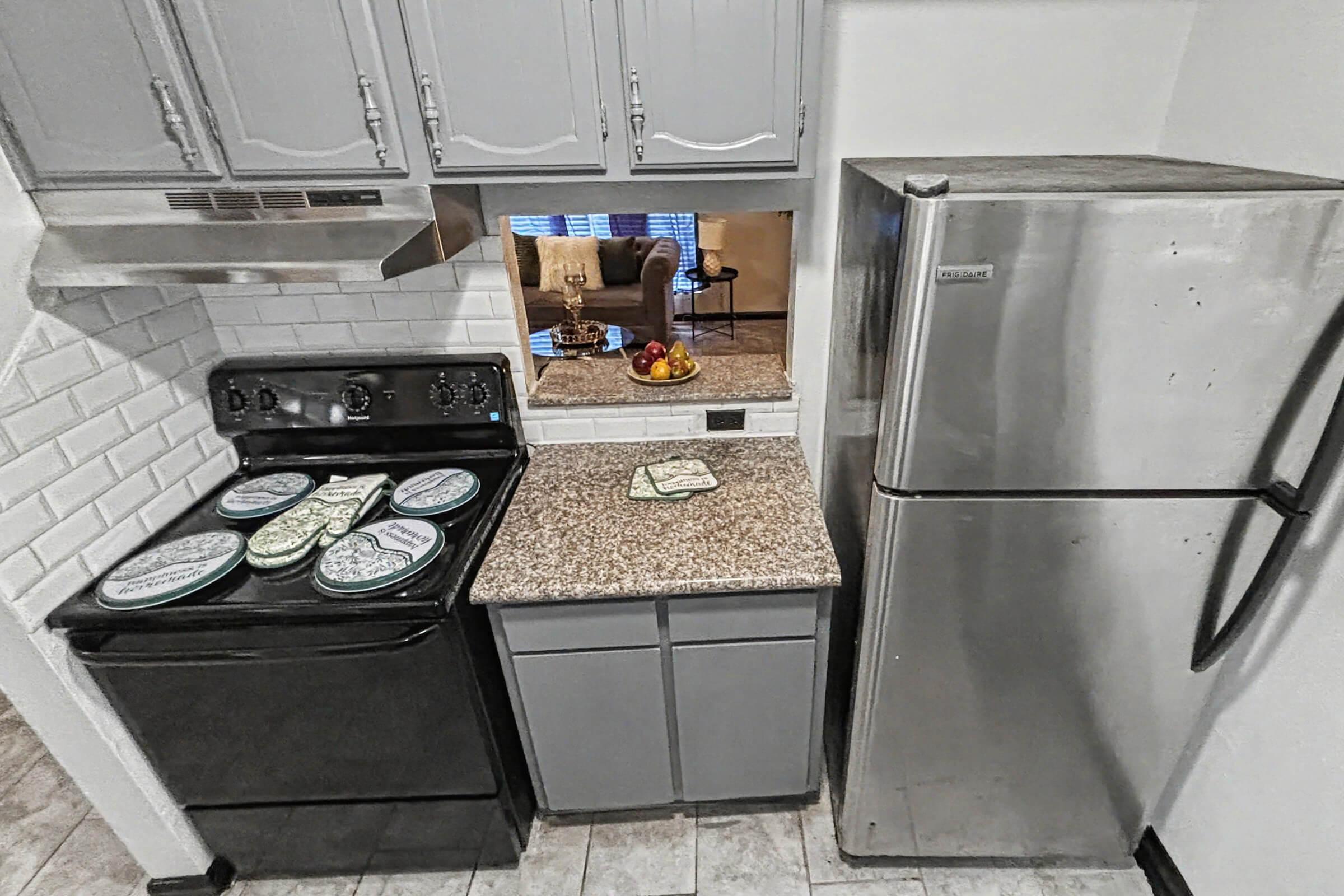 A compact kitchen featuring gray cabinets, a black stove, and a stainless steel refrigerator. There is a granite countertop with decorative plates arranged on it, and a small opening with a view of a living area through a window. The tiles on the floor are light-colored and provide contrast to the cabinetry.