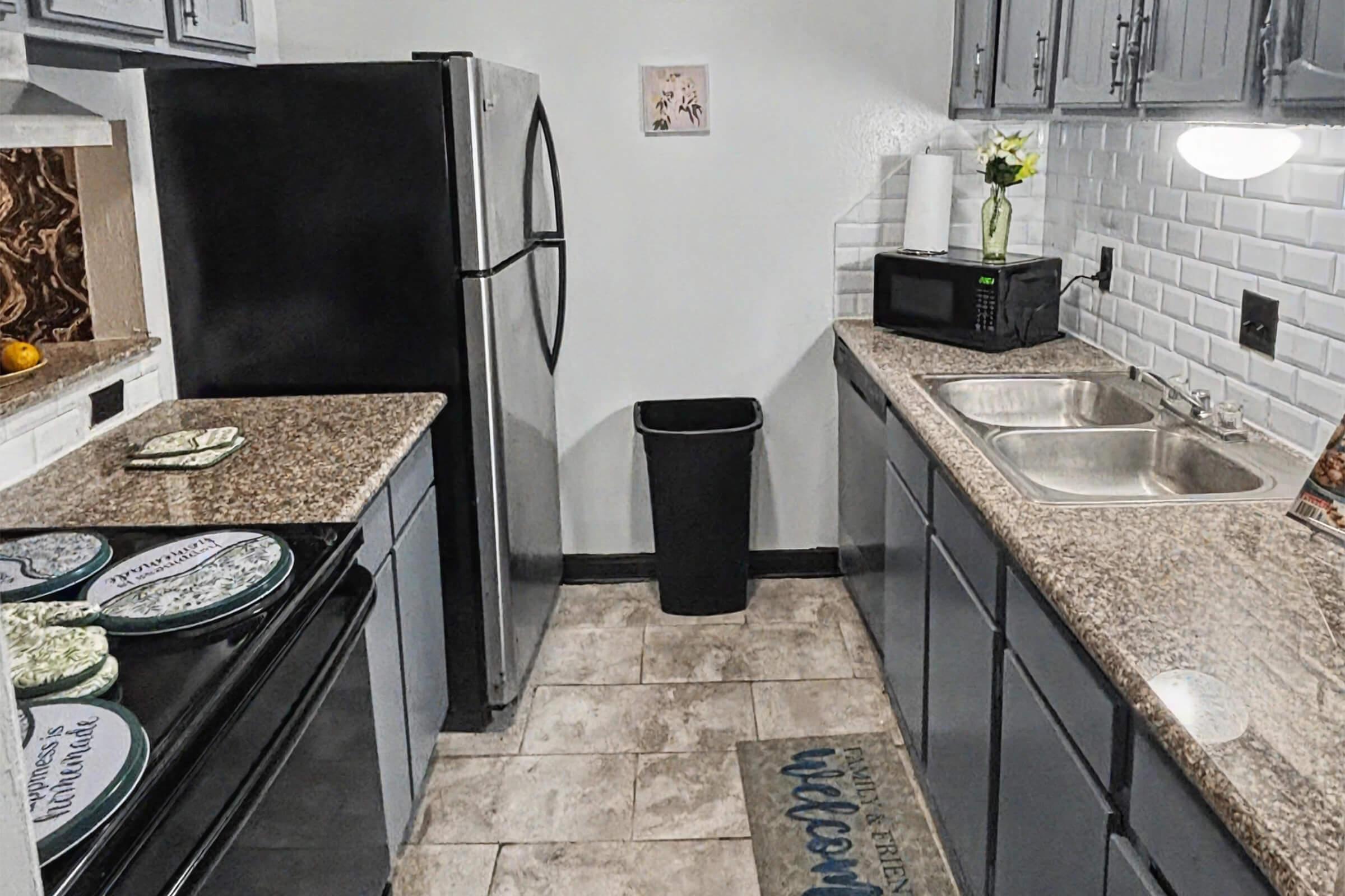 A modern kitchen featuring dark cabinets, a black refrigerator, and a sleek black microwave. The countertops are a light speckled granite. There are two sinks with chrome fixtures, a black trash bin, and decorative dishware. A small floral arrangement adds a touch of charm, while a "welcome" mat greets visitors.