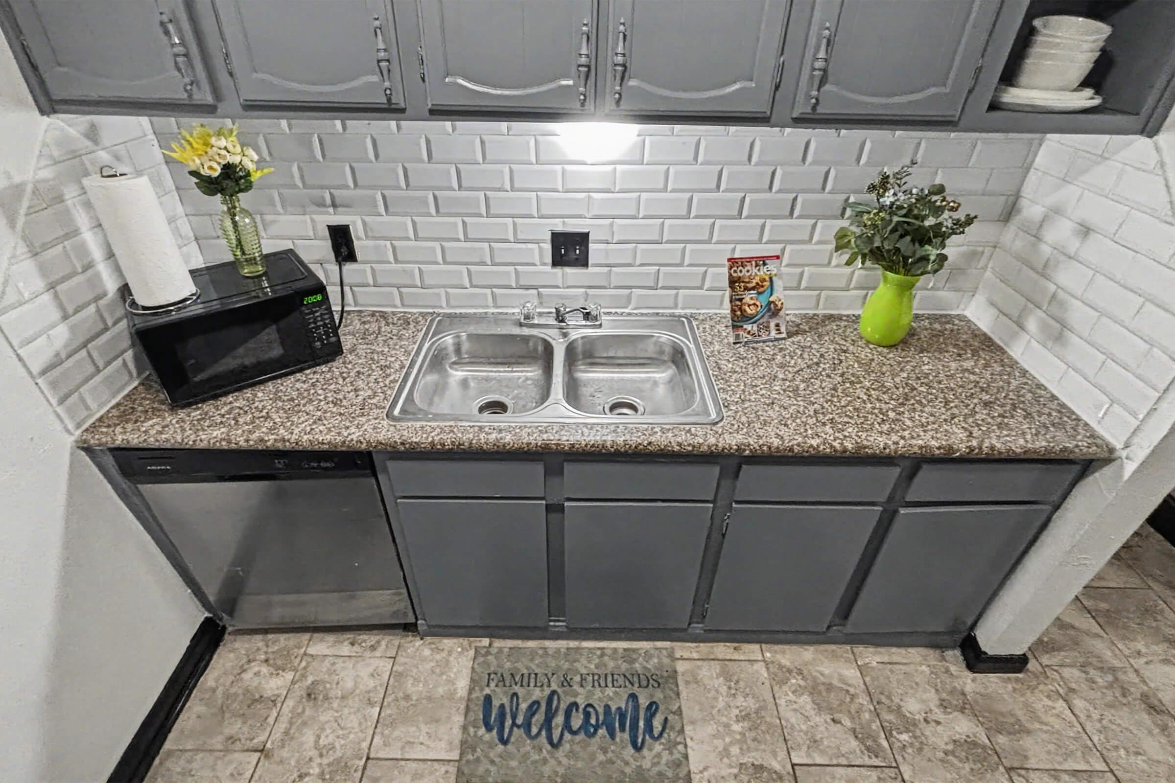 A tidy kitchen sink area featuring a granite countertop with a double sink, a microwave, and a dishwasher. Above the sink are cabinets, and there are decorative items including a vase with flowers and a box of cereal on the counter. The mat in front says "Family & Friends Welcome."