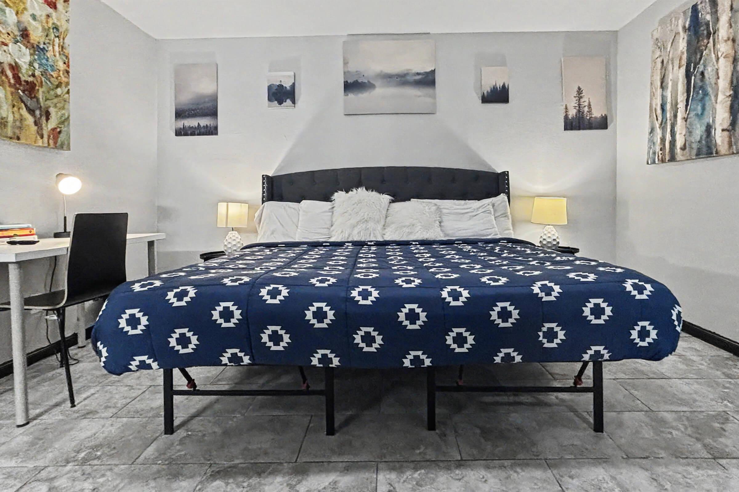 A cozy bedroom featuring a large bed with a blue patterned blanket, two bedside lamps, and various nature-themed artworks on the walls. There is a small desk and a black chair on one side, and the room has tiled flooring, creating a comfortable and inviting atmosphere.