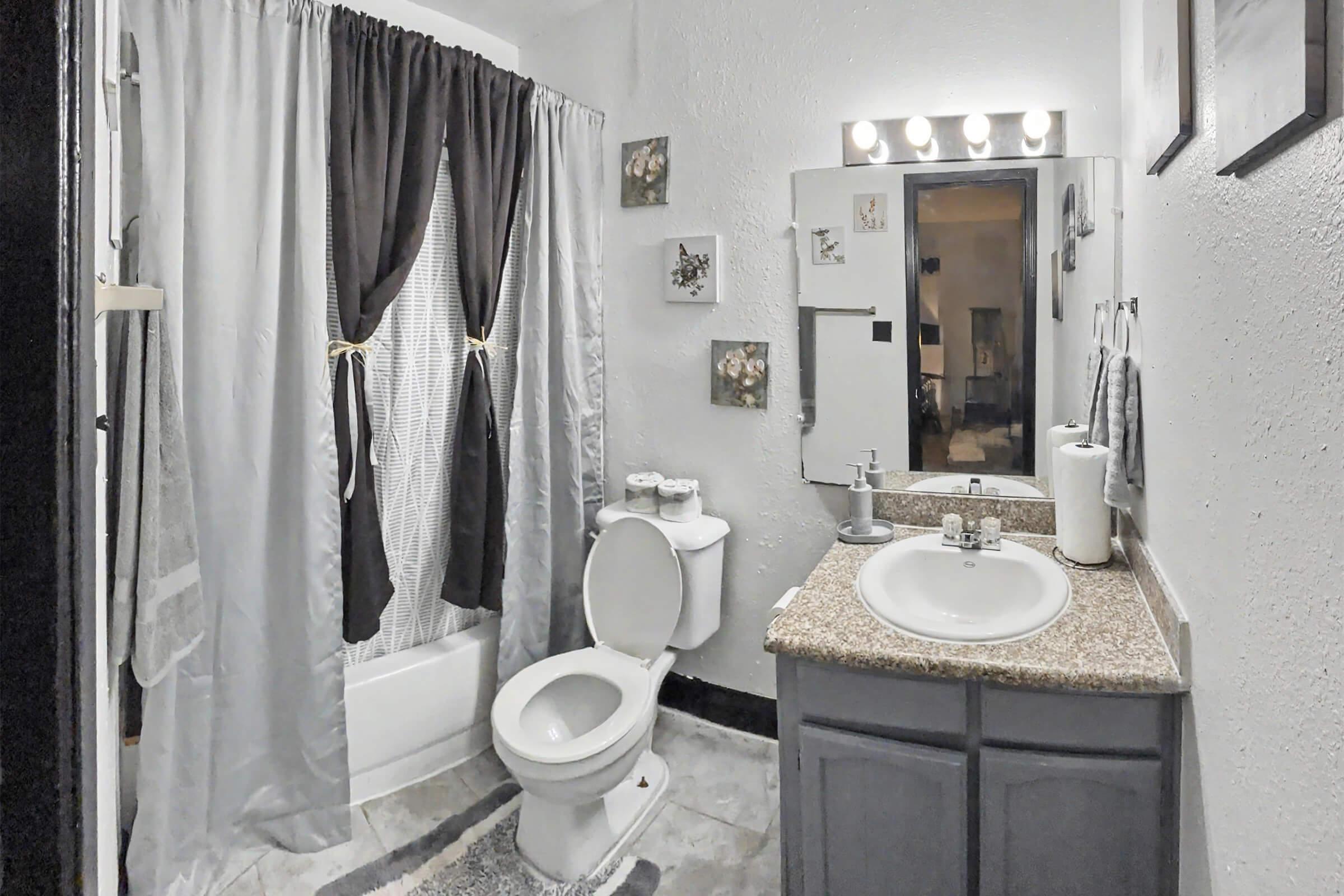 A small bathroom featuring a white toilet, a sink with a granite countertop, and a mirror above the sink. The shower area is adjacent, with a gray and white striped curtain. The walls are decorated with a few framed pictures, and the overall decor is simple and functional. Soft lighting enhances the warm ambiance.