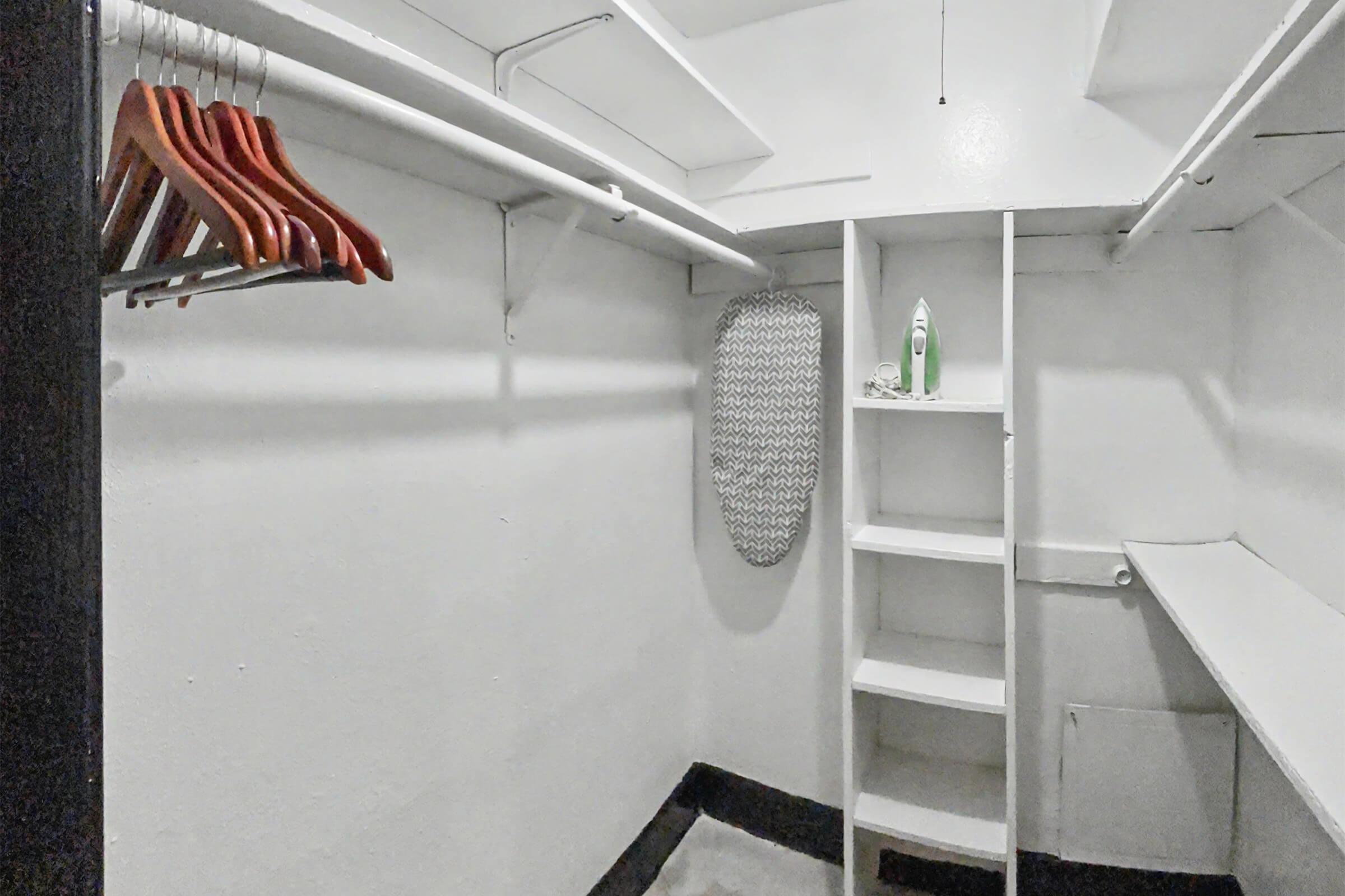 A spacious, empty closet featuring a row of wooden hangers on a white rod, a built-in shelving unit on one side, and a full-length mirror. The walls are painted white, and the floor is a light color, creating a clean and minimalist storage space.