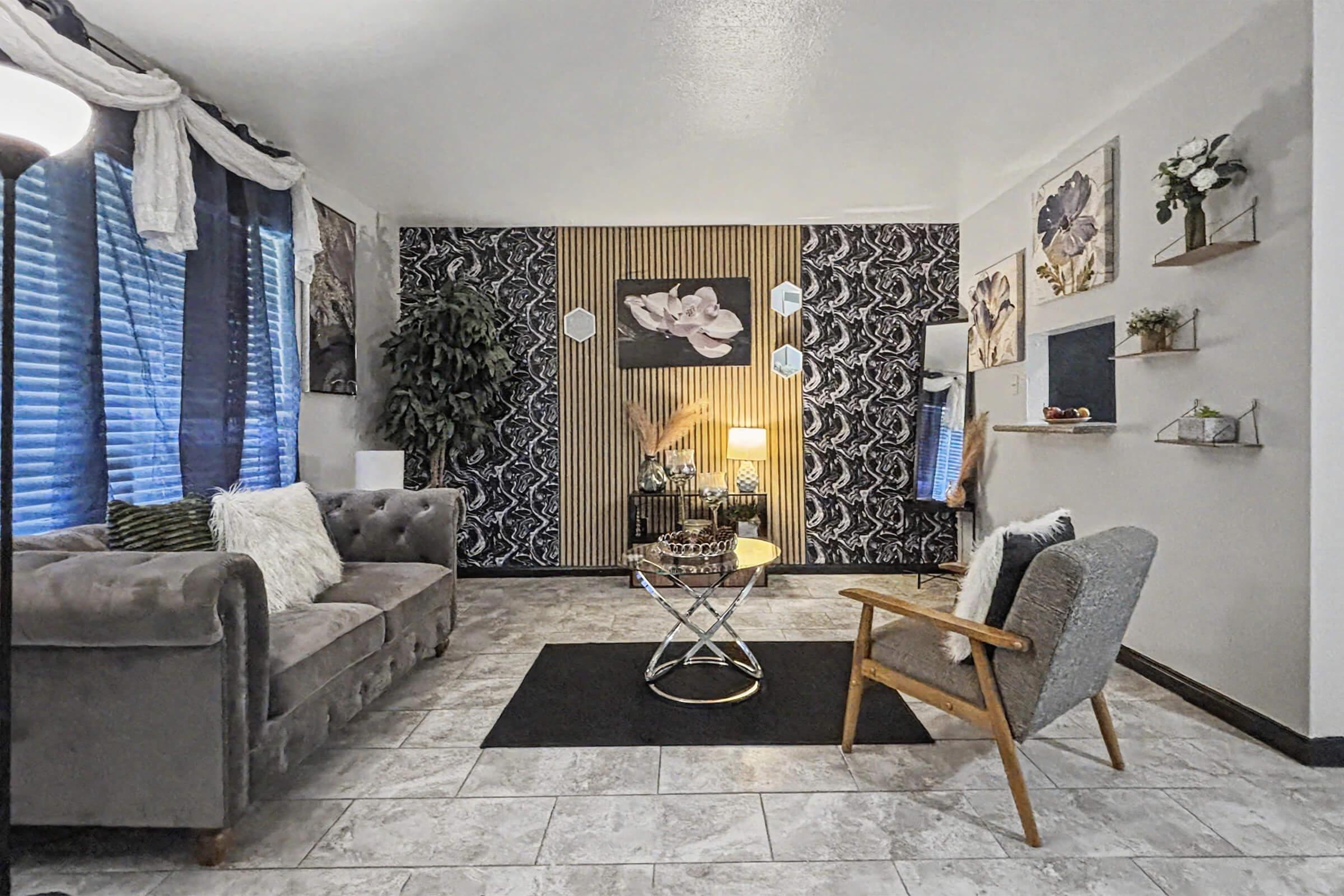 Cozy living room featuring a gray sofa, an armchair, and a glass coffee table. The walls are adorned with floral artwork and decorative shelves. Natural light filters through window treatments, illuminating a plant in the corner and a stylish rug on the tiled floor, creating a welcoming atmosphere.